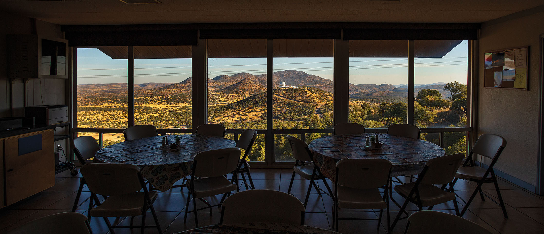 Large windows overlooking mountains and a telescope are at the back of a dimly-lit dining room with circular tables