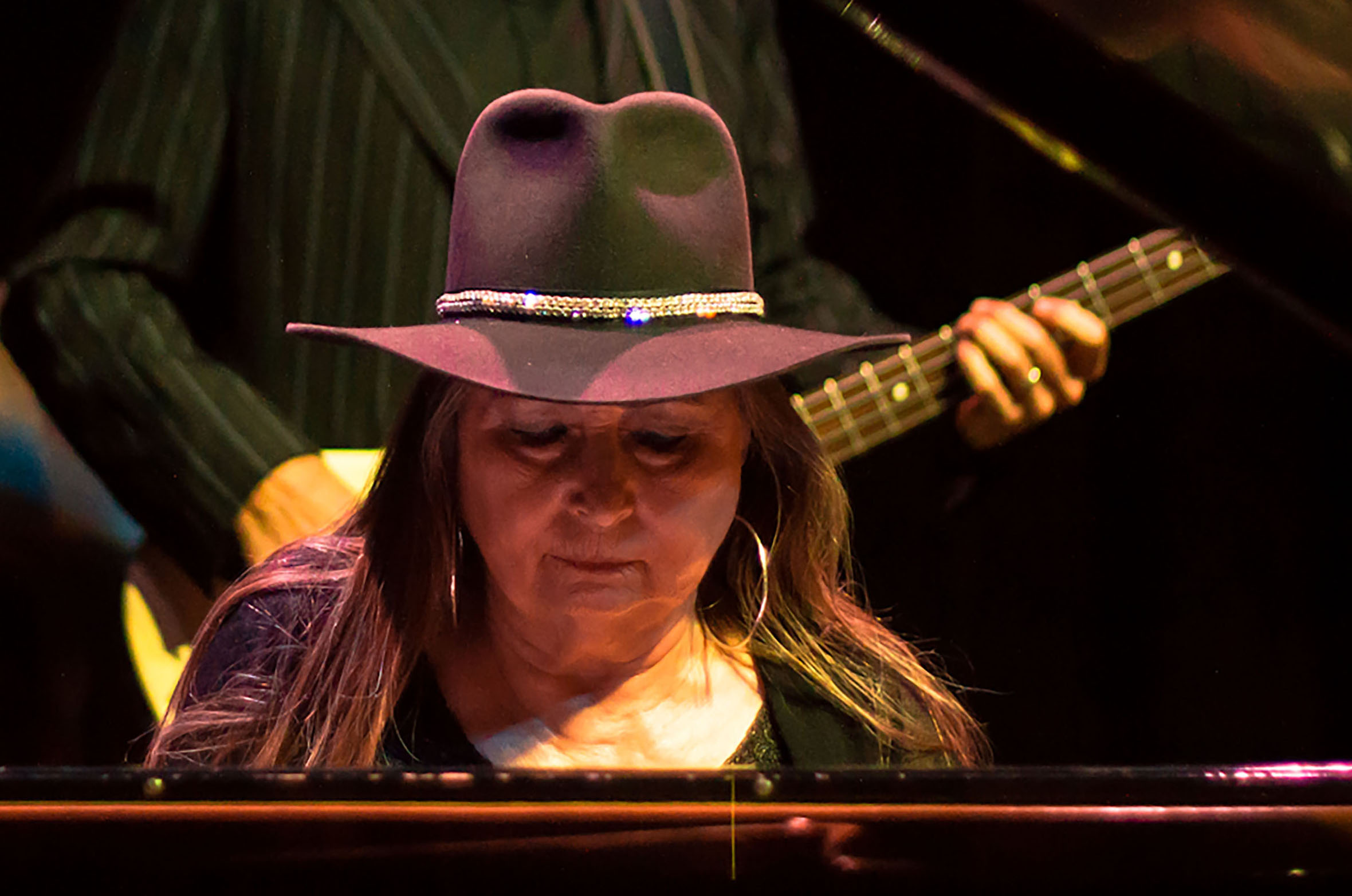 Bobbie Nelson, wearing a sequined black hat, plays the piano in front of a band
