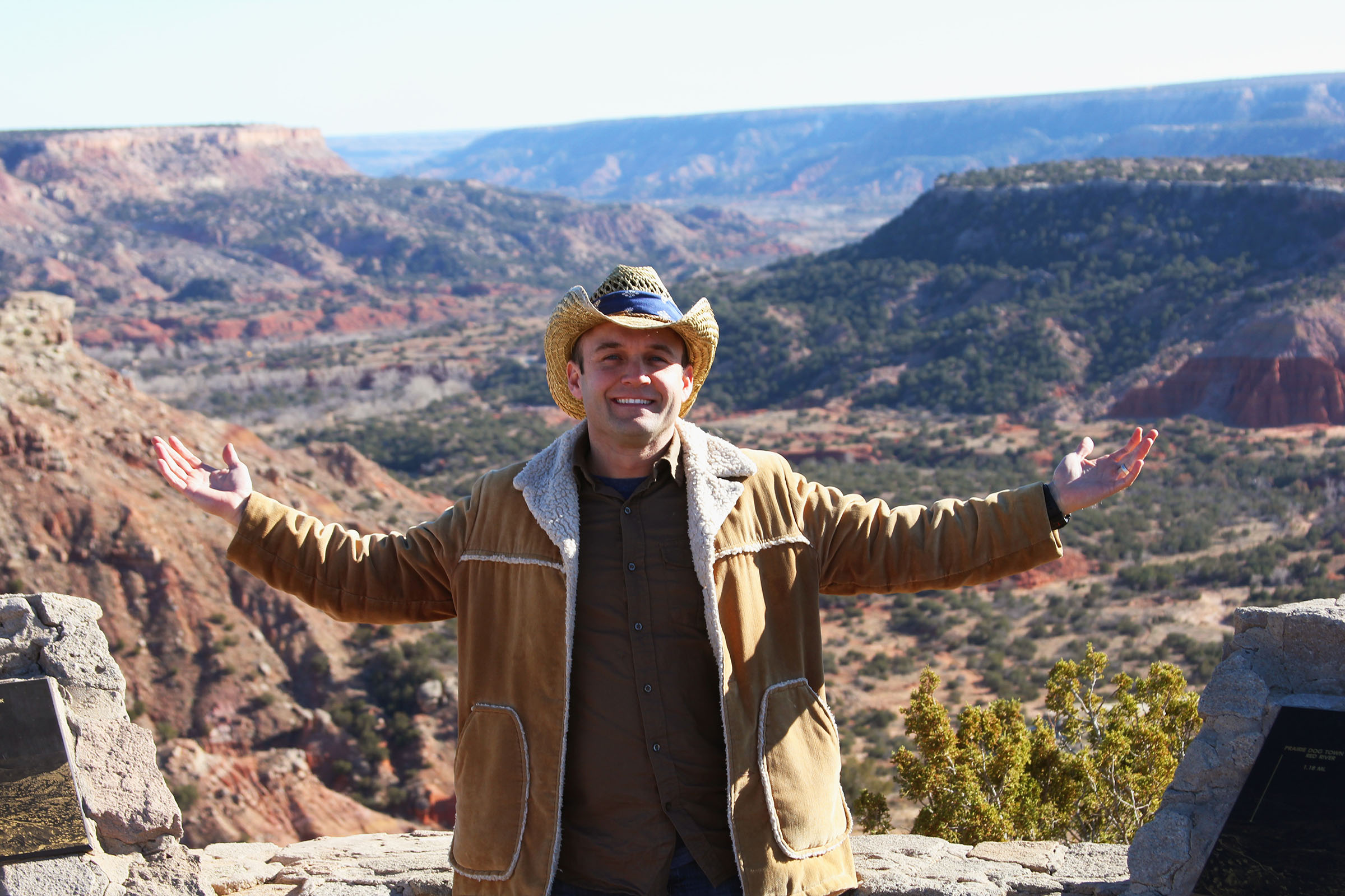 A picture of a man wearing a tan jacket holding his arms up in the air, in front of a large expanse of canyonlands