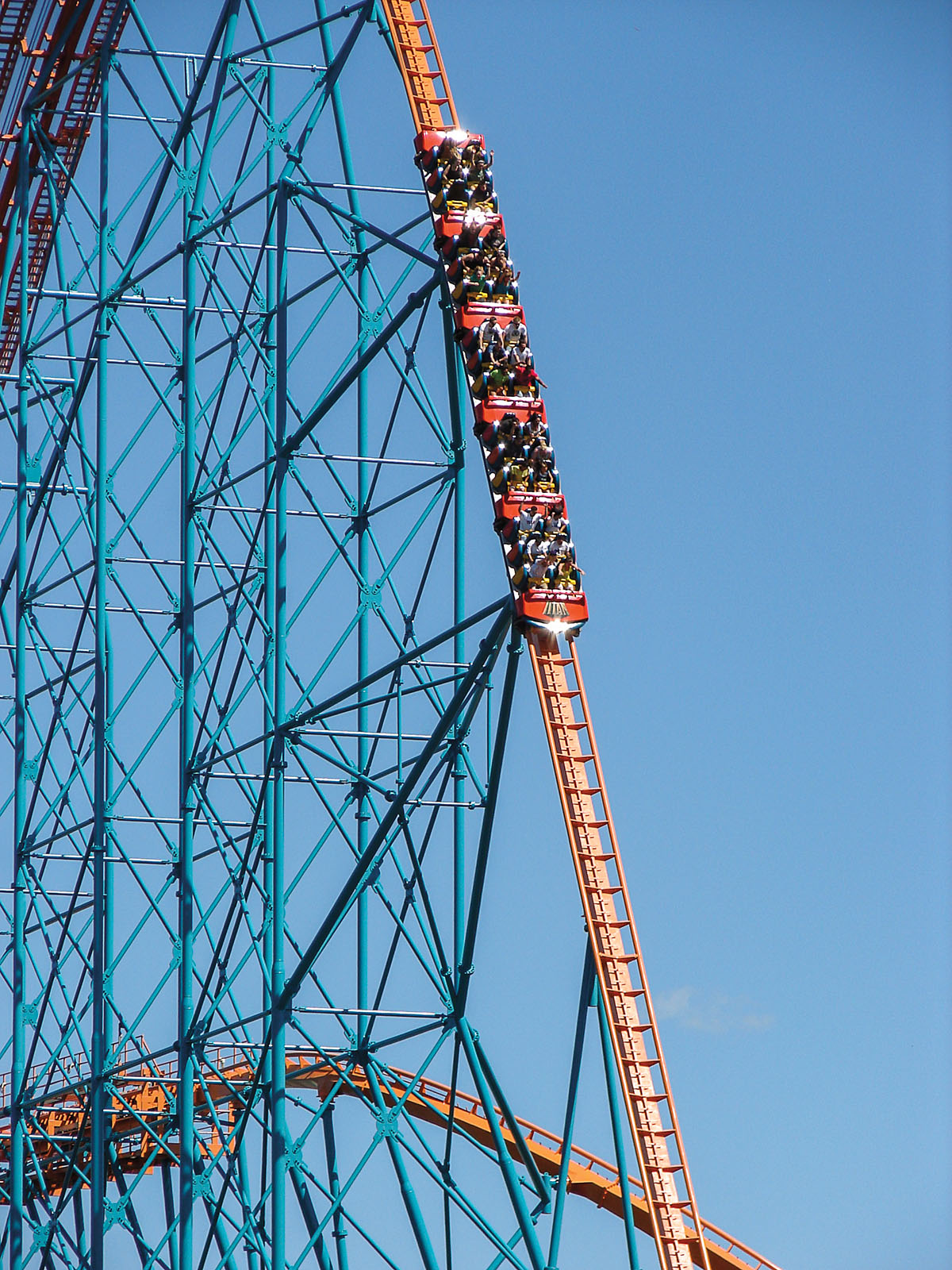 A photo of a very steep roller coaster with small red cars on blue sky