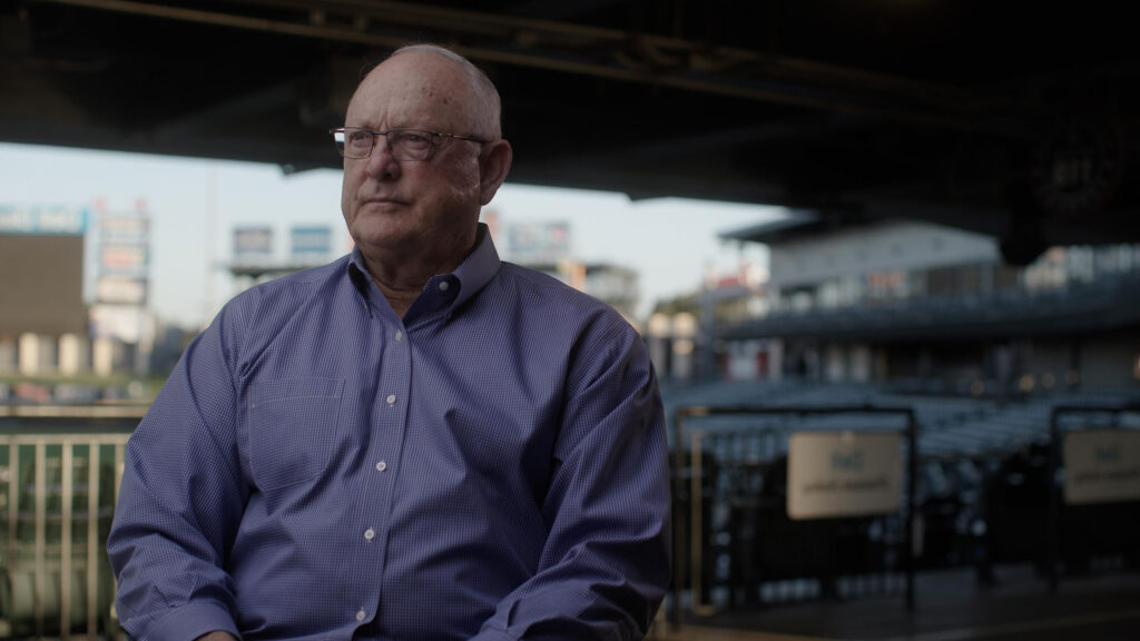 A man in a blue button-down shirt stares just off from the camera inside of a baseball stadium