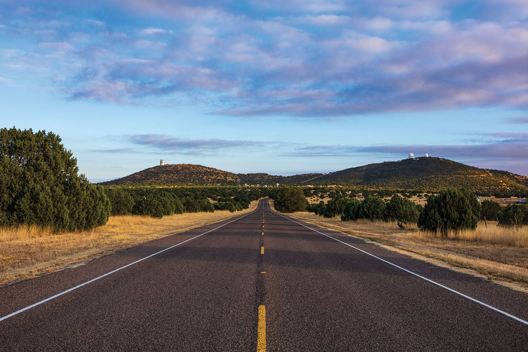 A photo of a wide open highway leading toward two mountains under a blue sky with clouds