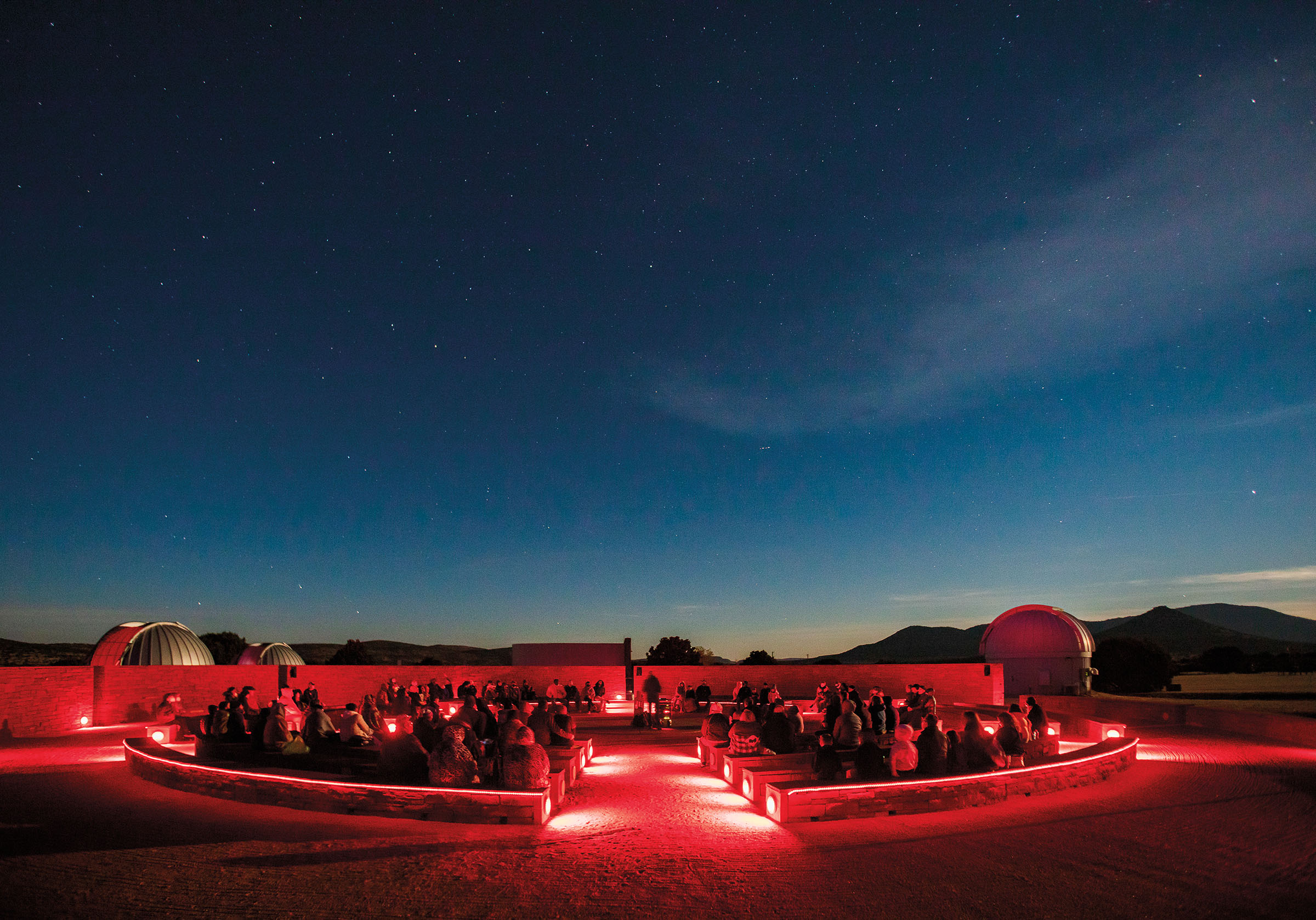 A low red glow illuminates an audience of spectators underneath a dark night sky full of stars
