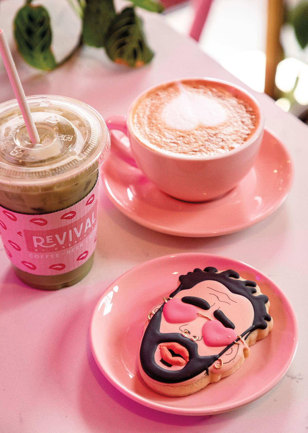 A picture of a latte in a pink ceramic cup, an iced coffee, and a sugar cookie of a man's face wearing pink sunglasses
