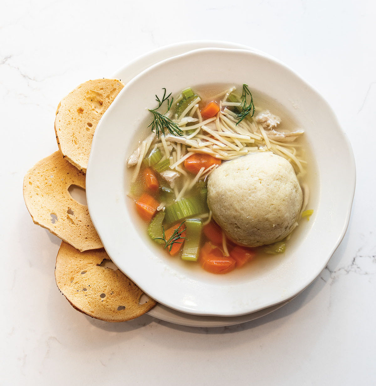 An overhead picture of a bowl of matzo ball soup with celery and carrots, accompanied by small crackers