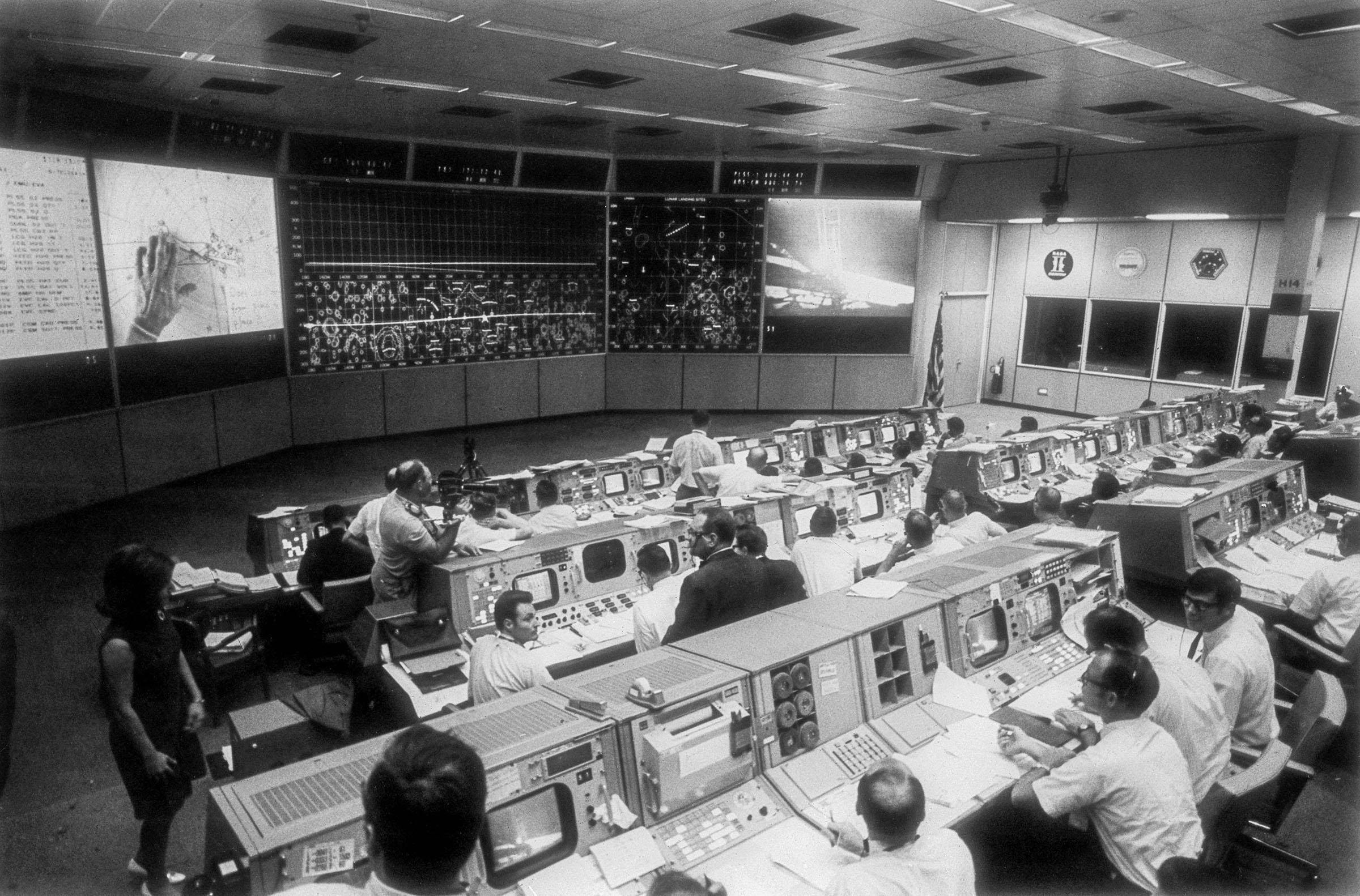 A historical black and white photgraph of people working in a large control rooom with several computer monitors and screens