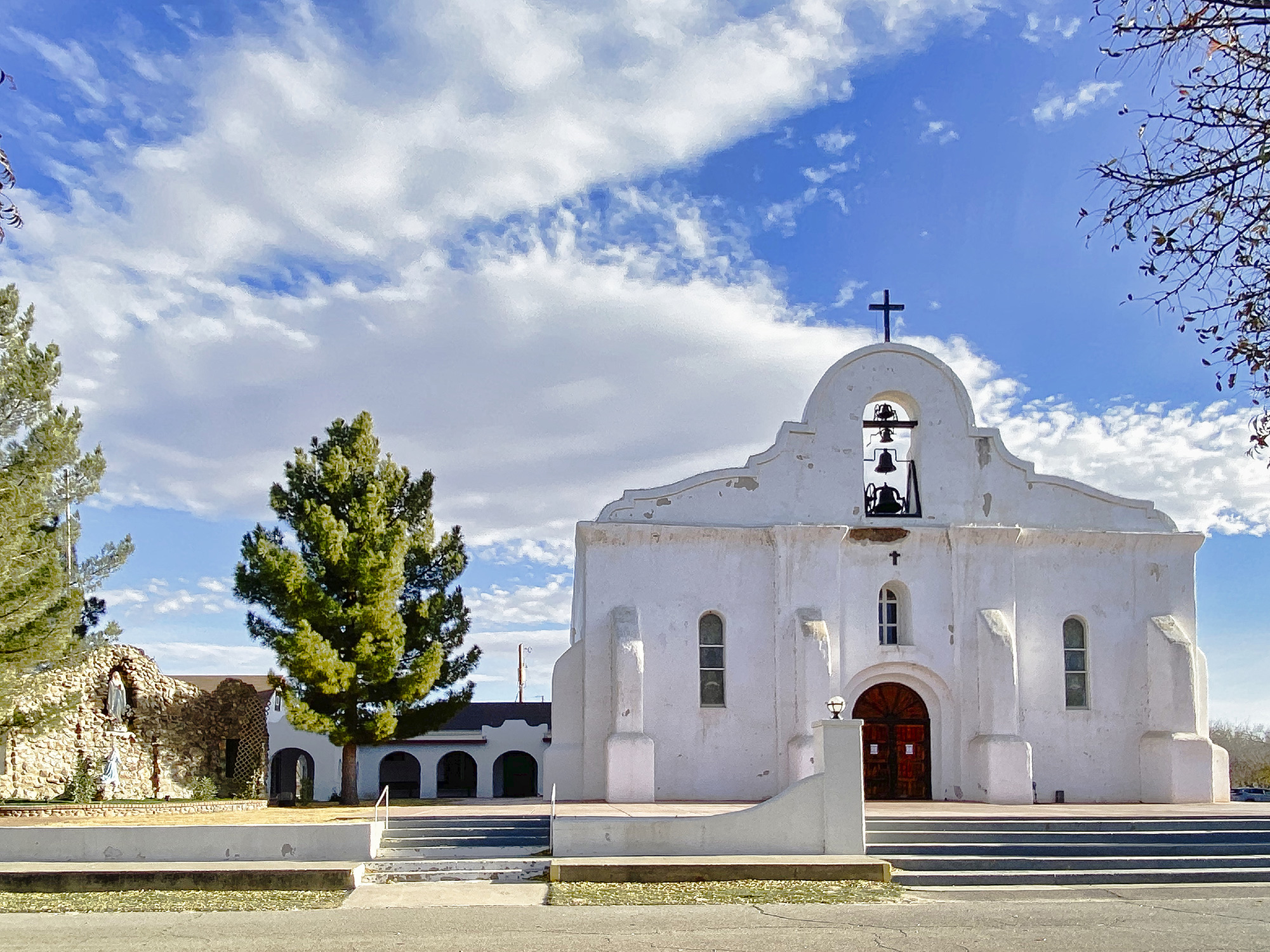 The San Elizario presidio chapel stands next to tall trees and a sunny but partly cloudy sky