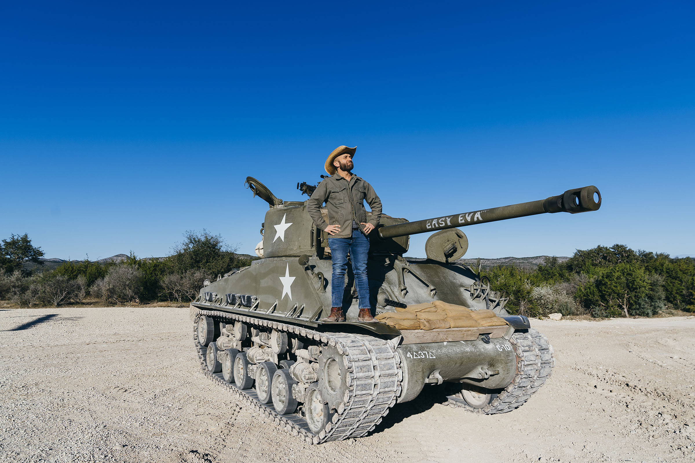 A man in a cowboy hat stands on top of a large green tank on dusty ground