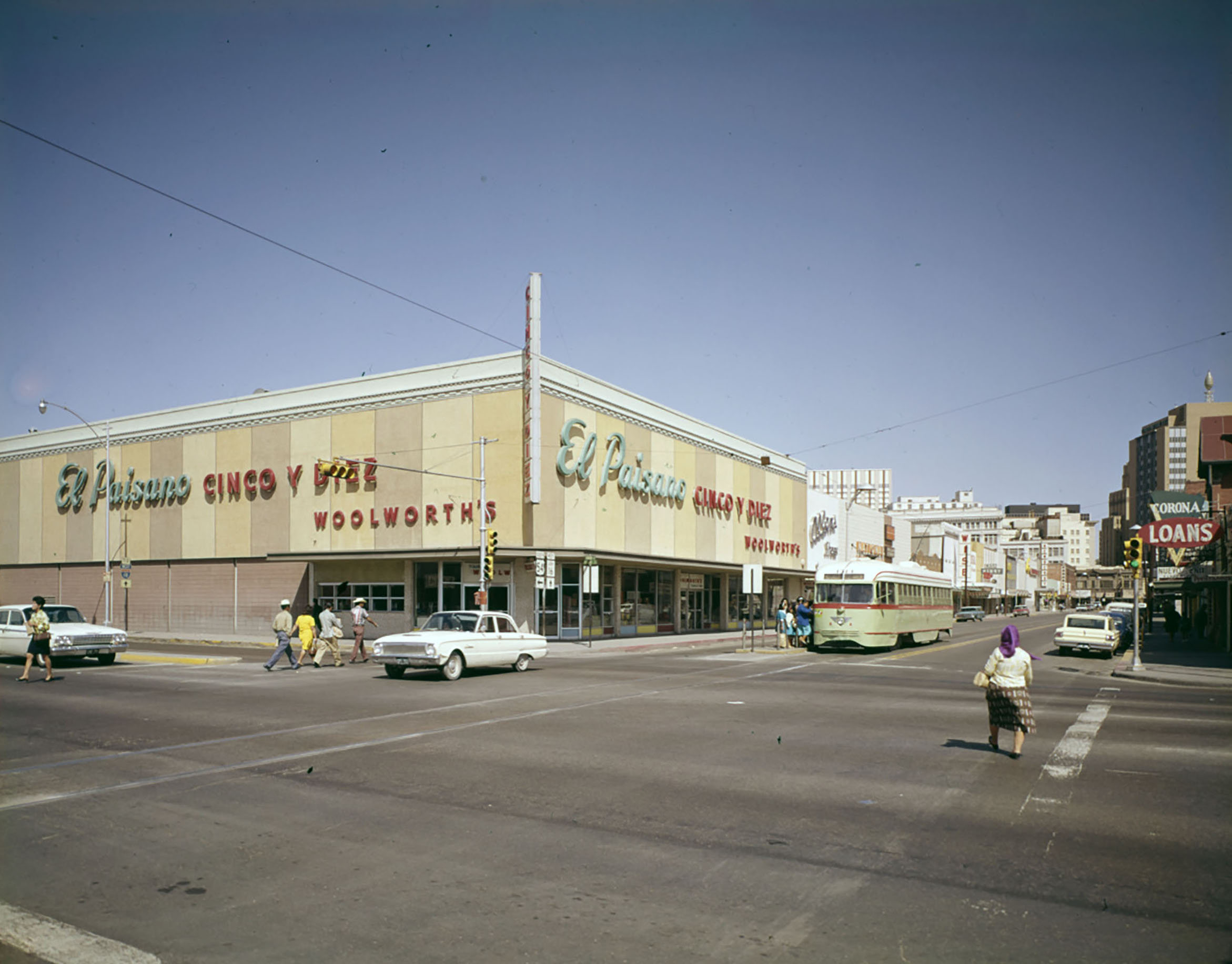 A view of a large store on a city street. A streetcar and pedestrian pass by large 1960s automobiles