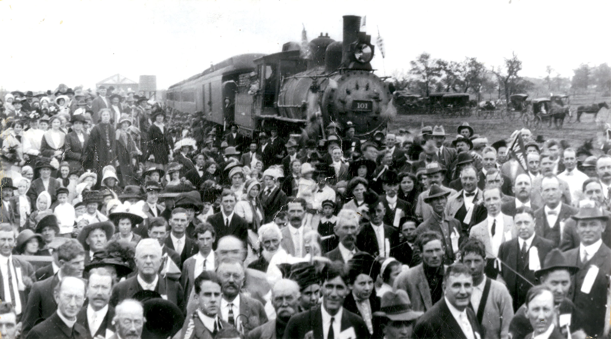 A large crowd of people, dressed in suits, ties and hats, stand in front of a locomotive in this black and white photograph