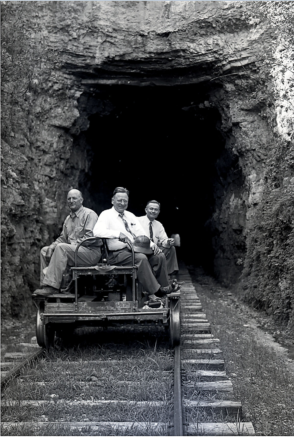 Three men in button-down shirts ride on a hand cart on train tracks going into a stone tunnel