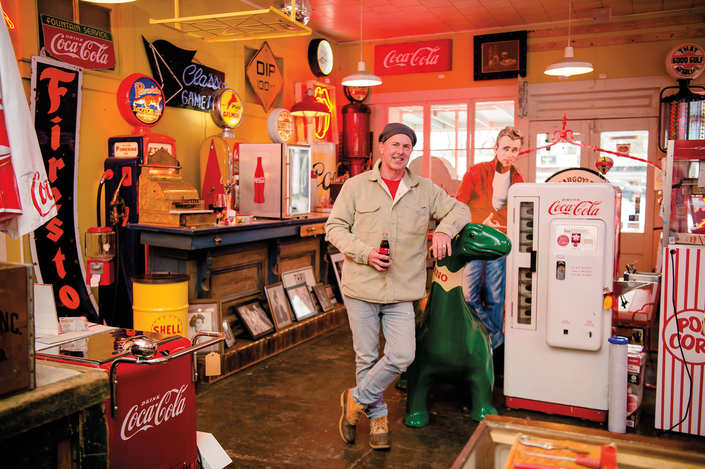A man holds a glass-bottled Coca Cola in his hand and leans against a green dinosaur, in a room filled with vintage memorabilia
