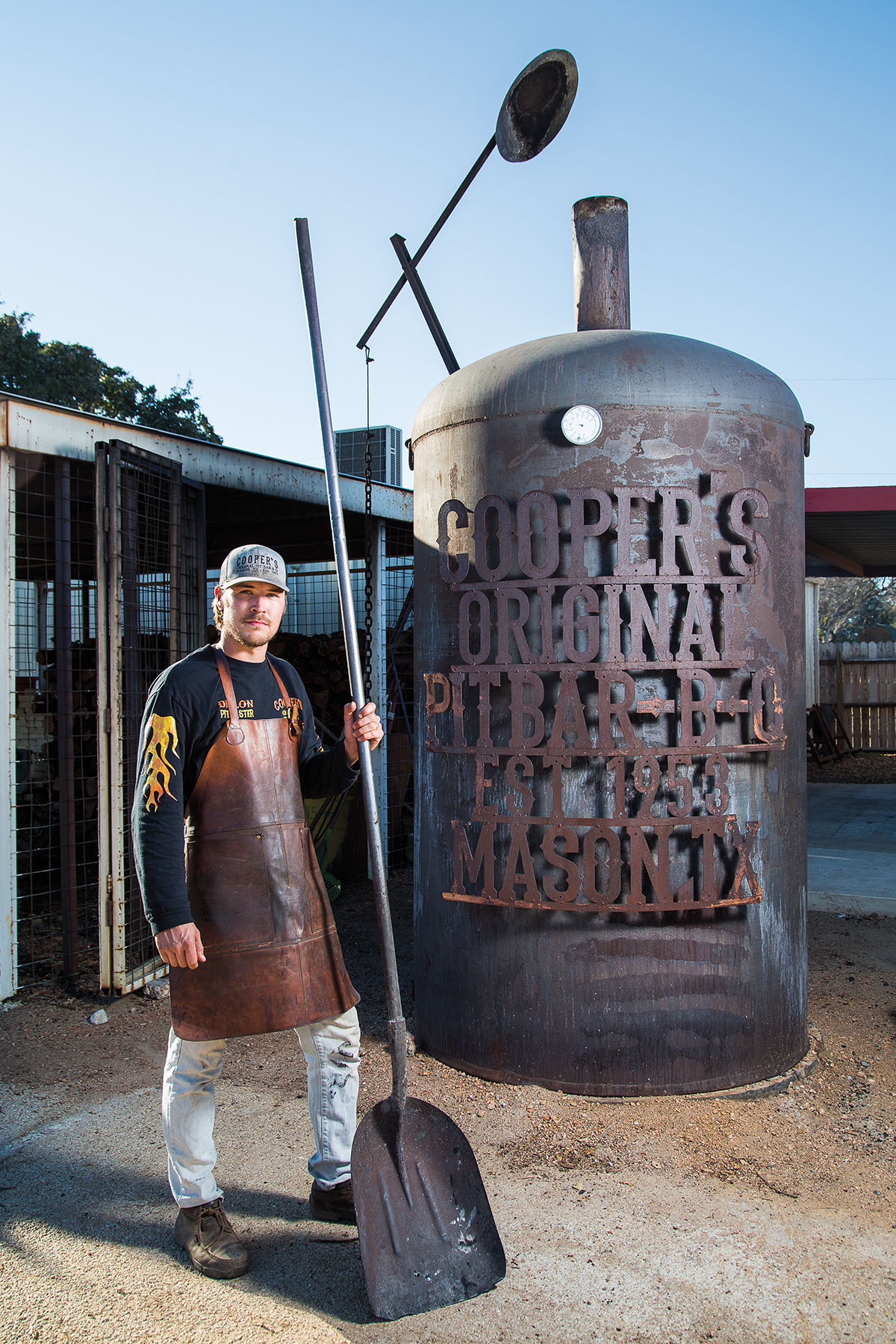 A man in a baseball cap stands in front of a large metal smoker reading "Cooper's Original Pit BBQ"