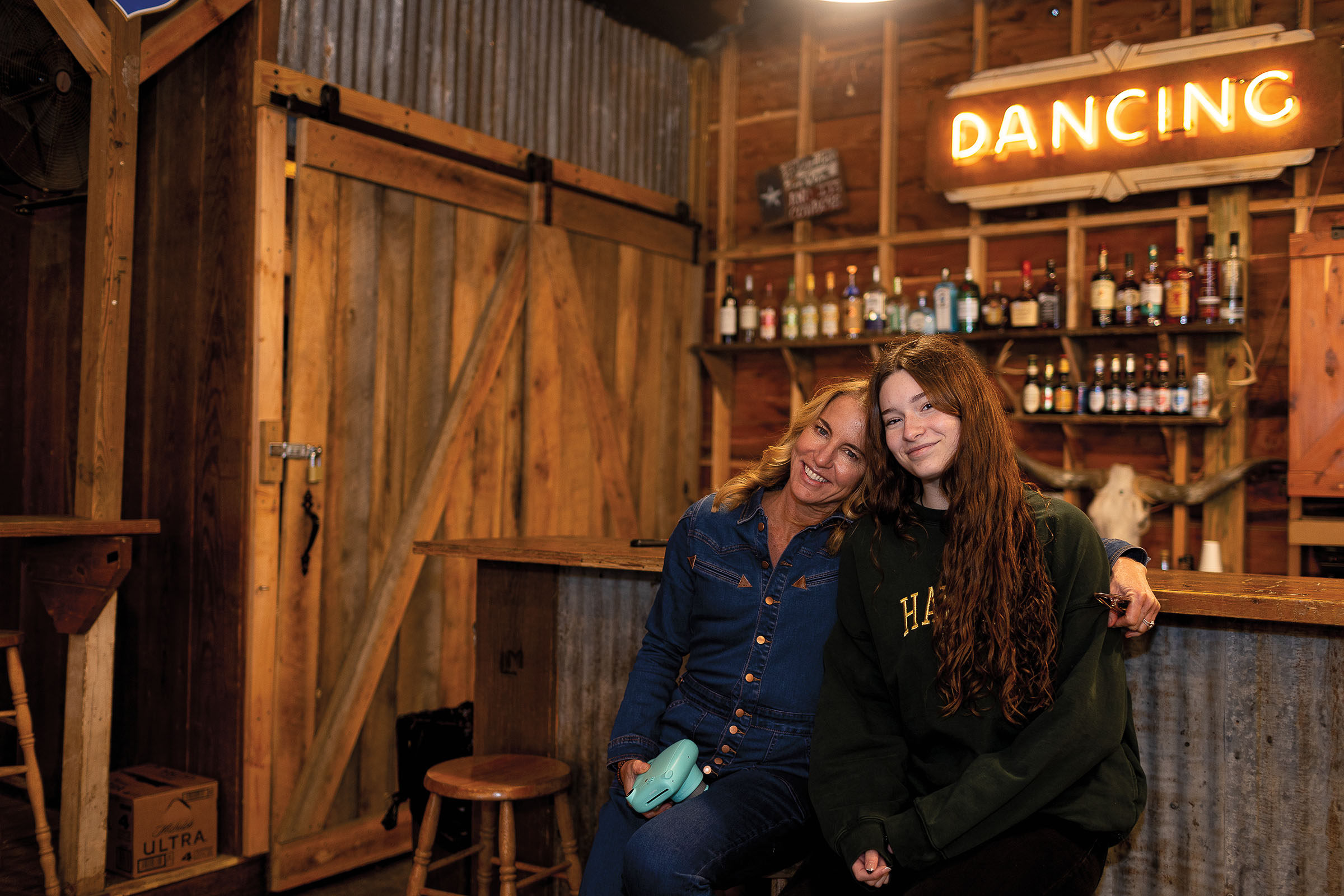 A woman and young woman lean together in front of a neon sign reading "Dancing"