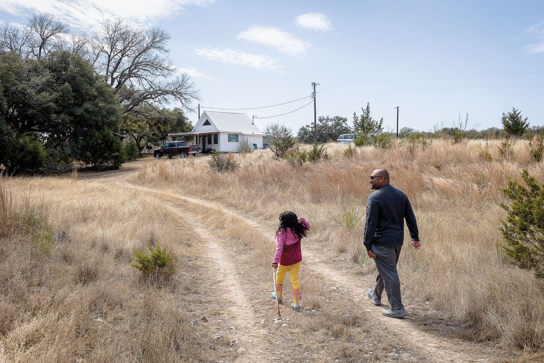 A man and his daughter walk down a tire-track dirt path in a dry Hill Country landscape with a white house in the background