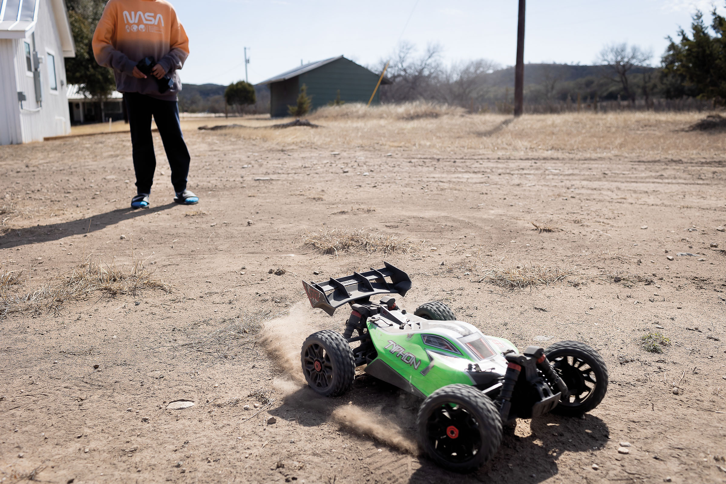 A young man wearing a shirt reading "NASA" drives a bright green remote controlled car on a dirt path