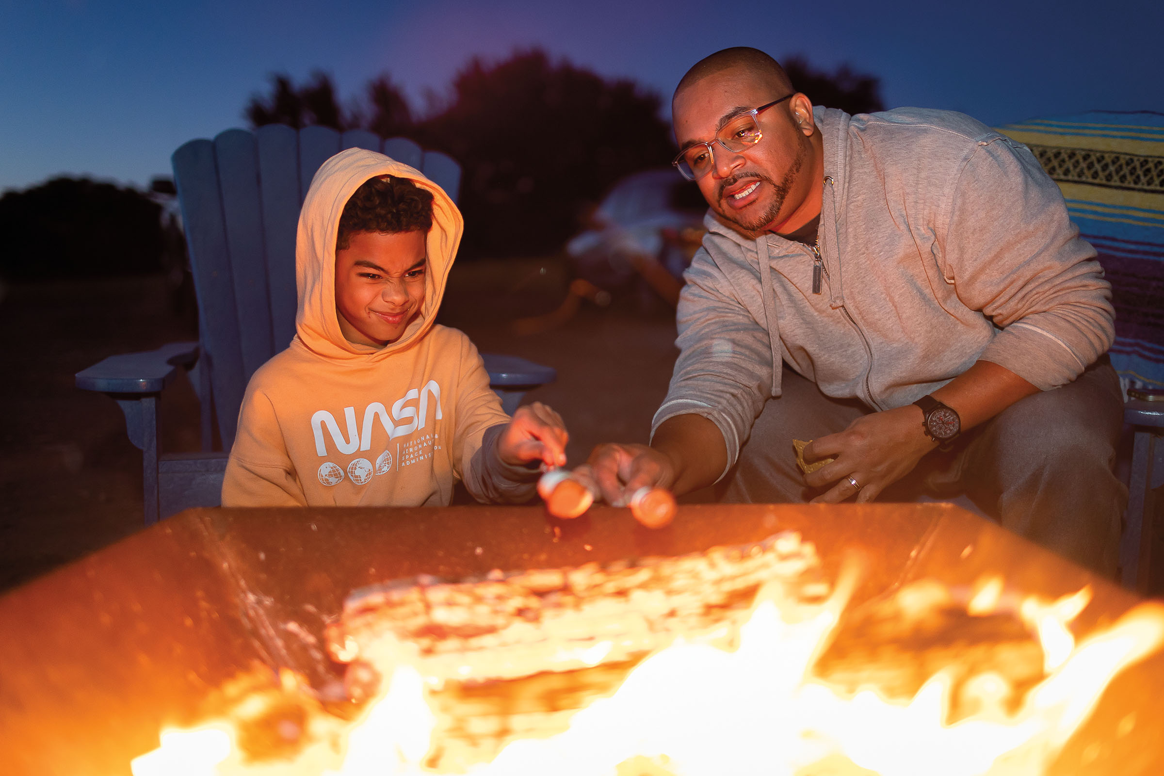 A young man and his father hold sticks with toasted marshmallows in front of a roaring fire
