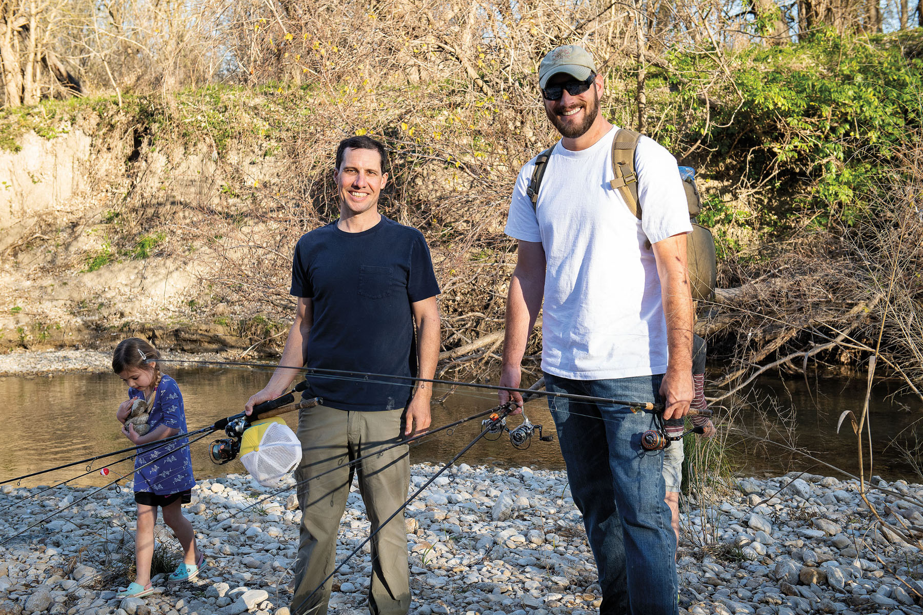 A young person and two adult men stand in t-shirts holding fishing rods along a rocky embankment