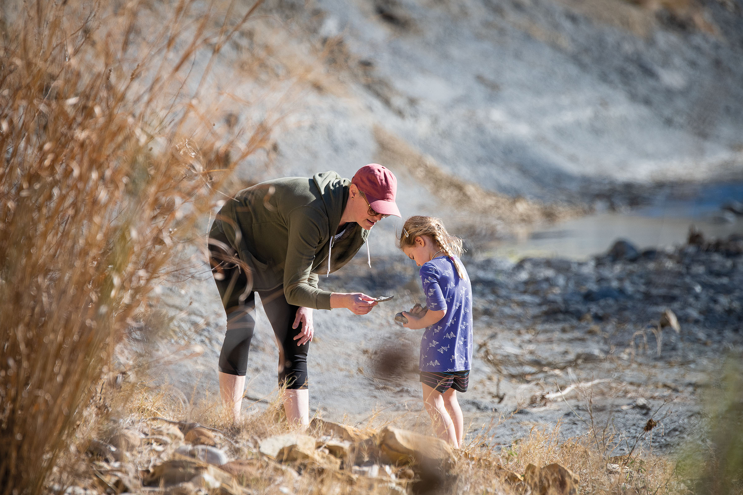 A person leans down to show their daughter a rock in a gray and rocky area of a riverbed