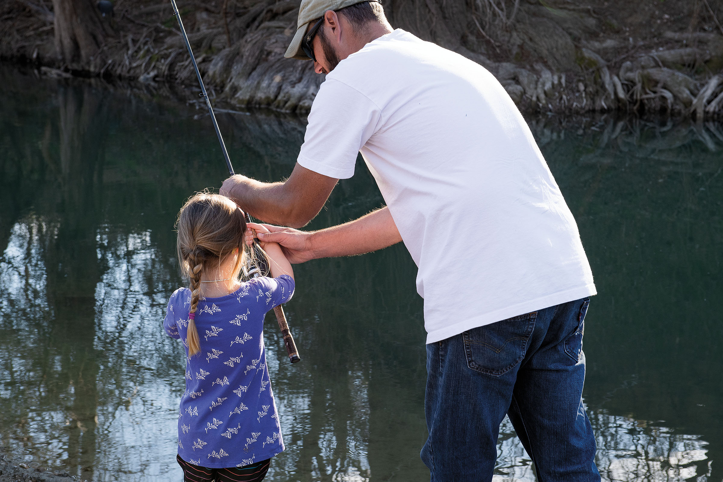 A man helps a young woman with a fishing rod next to a creek