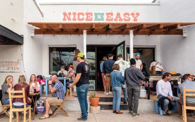 The exterior of a whitewashed building reading "Nice N Easy" where people gather and sit on wooden chairs