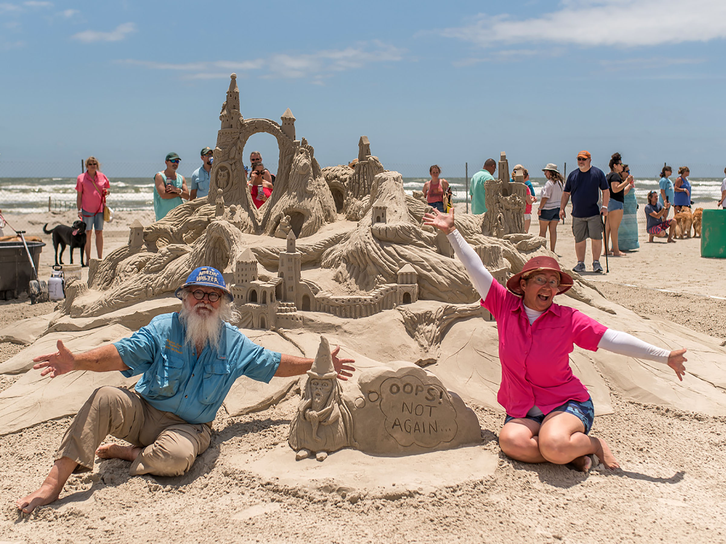 A picture of two people sitting in the sand with their arms outstretched in front of an elaborate sand castle design with gnomes, arches, pathways and signs