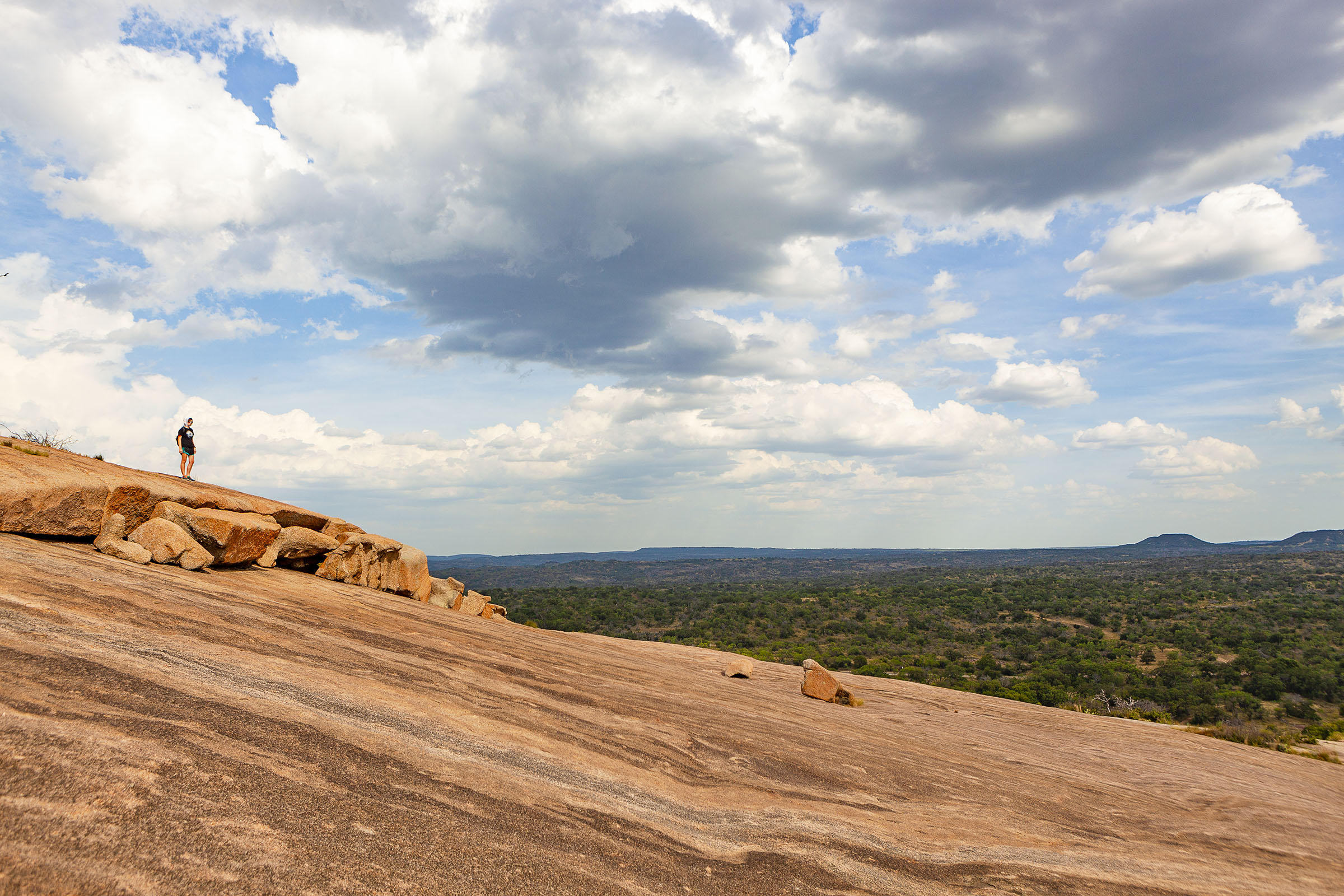A wide view of the red-gray surface of Enchanted Rock, high in the air against a green backdrop under blue sky