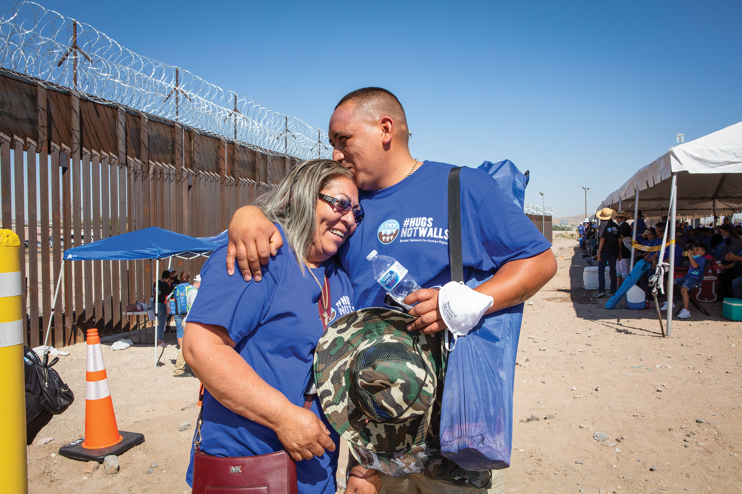 Two peopleembrace and kiss wearing blue shirts in front of a large steel border wall