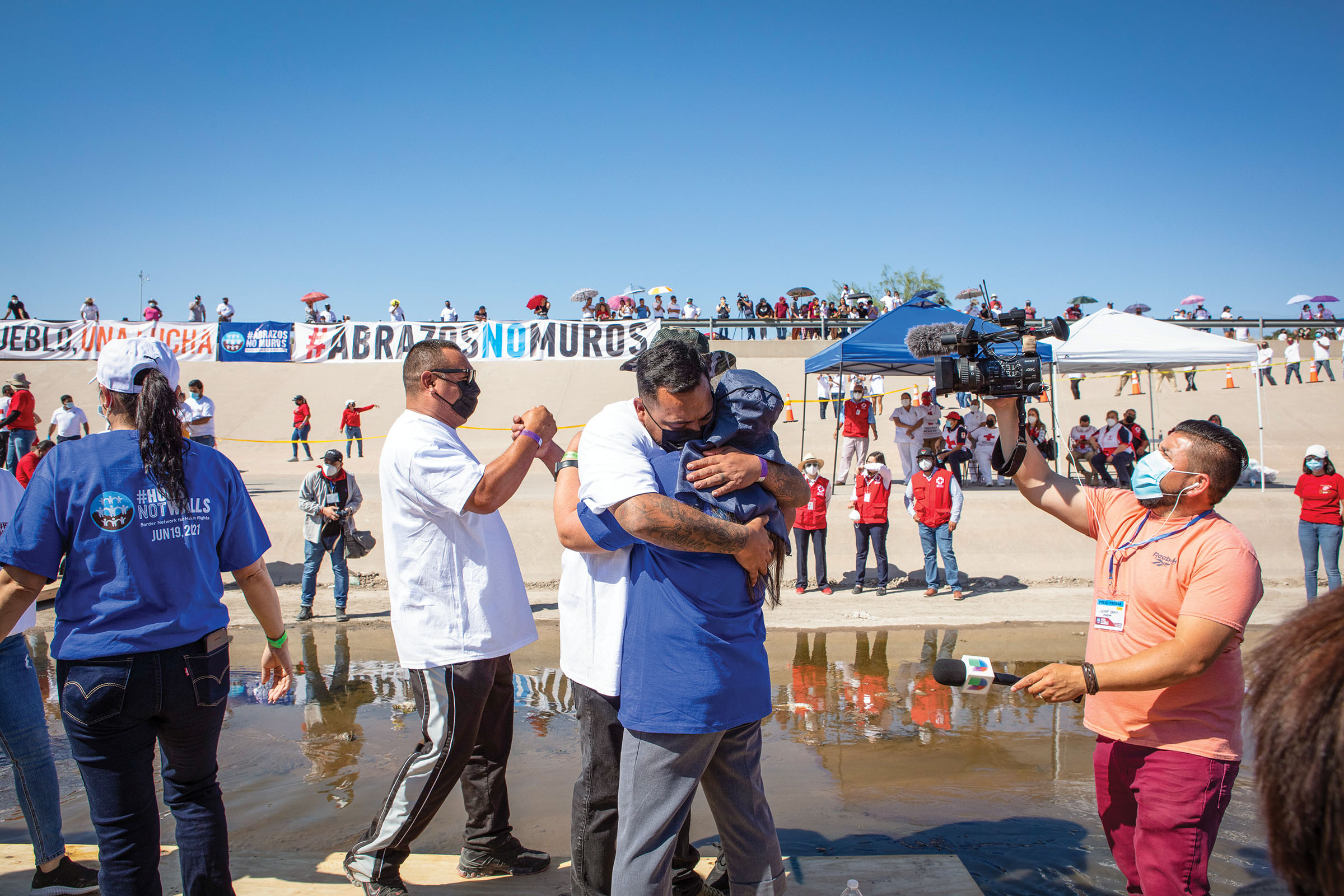 Two people embrace while others move around in the background offering fist bumps under blue sky along the Rio Grande
