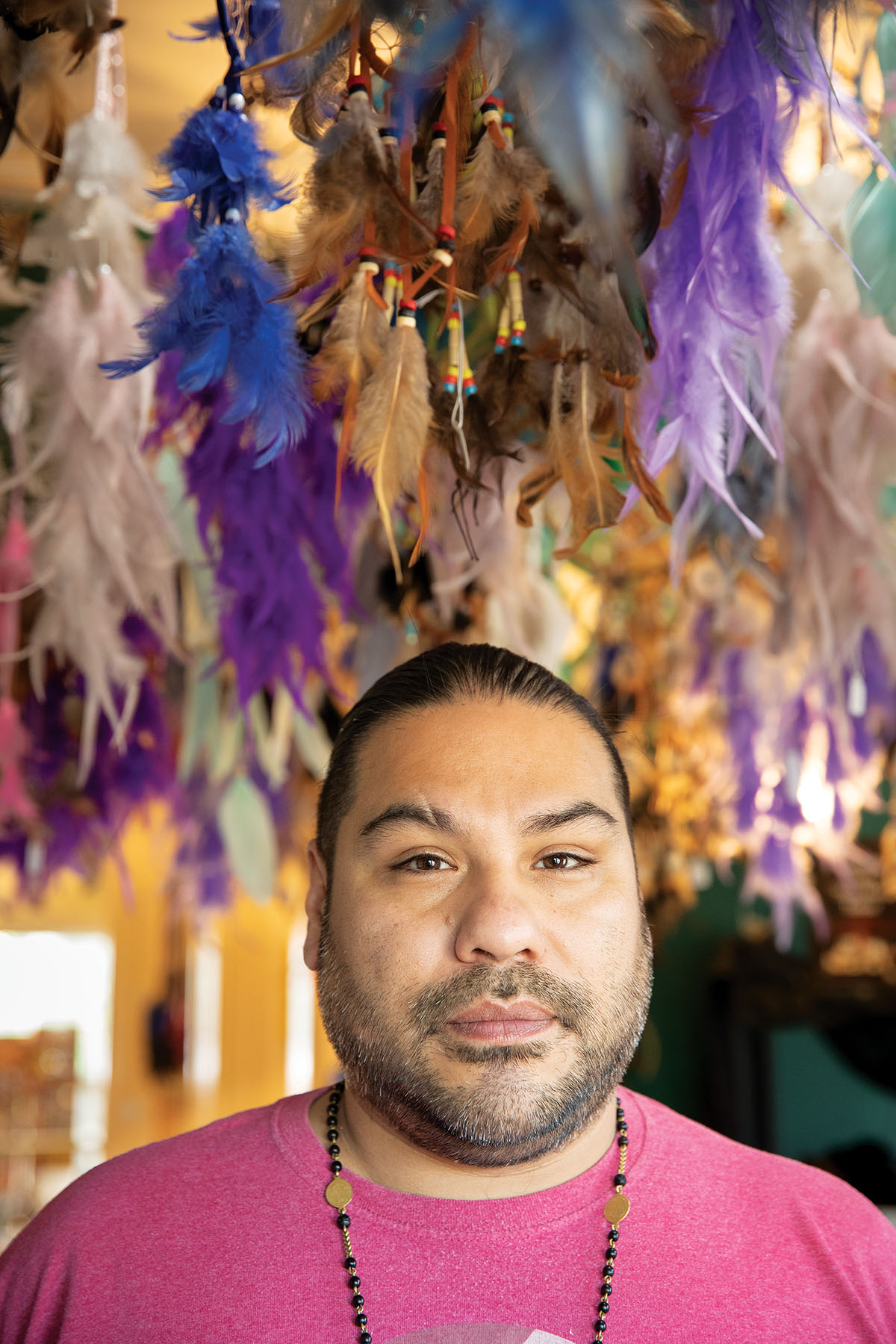 A man in a purple shirt stands in front of a brightly colored background with numerous feathers