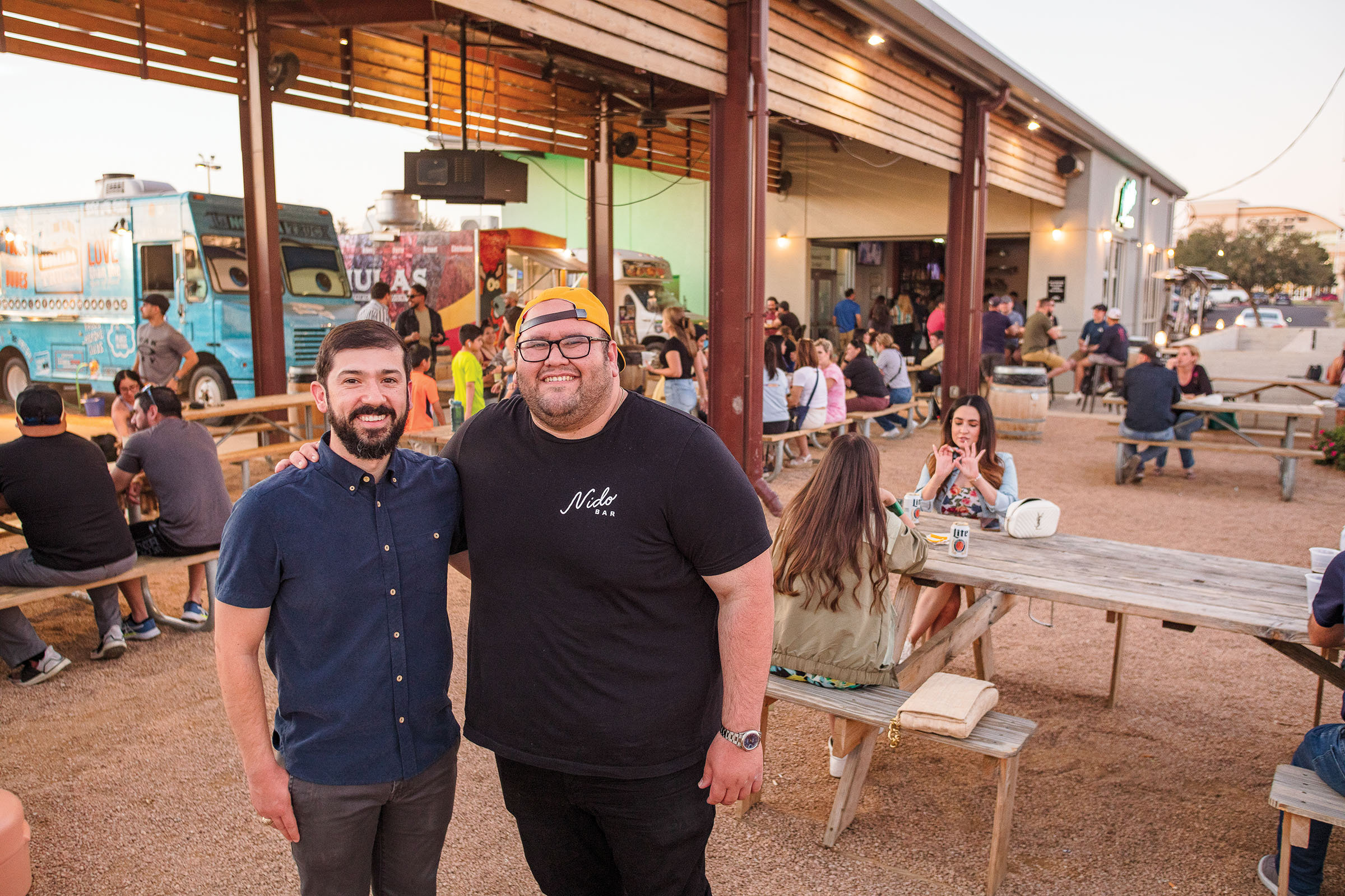 Two men stand in a dirt-floored food court with several food trucks nearby
