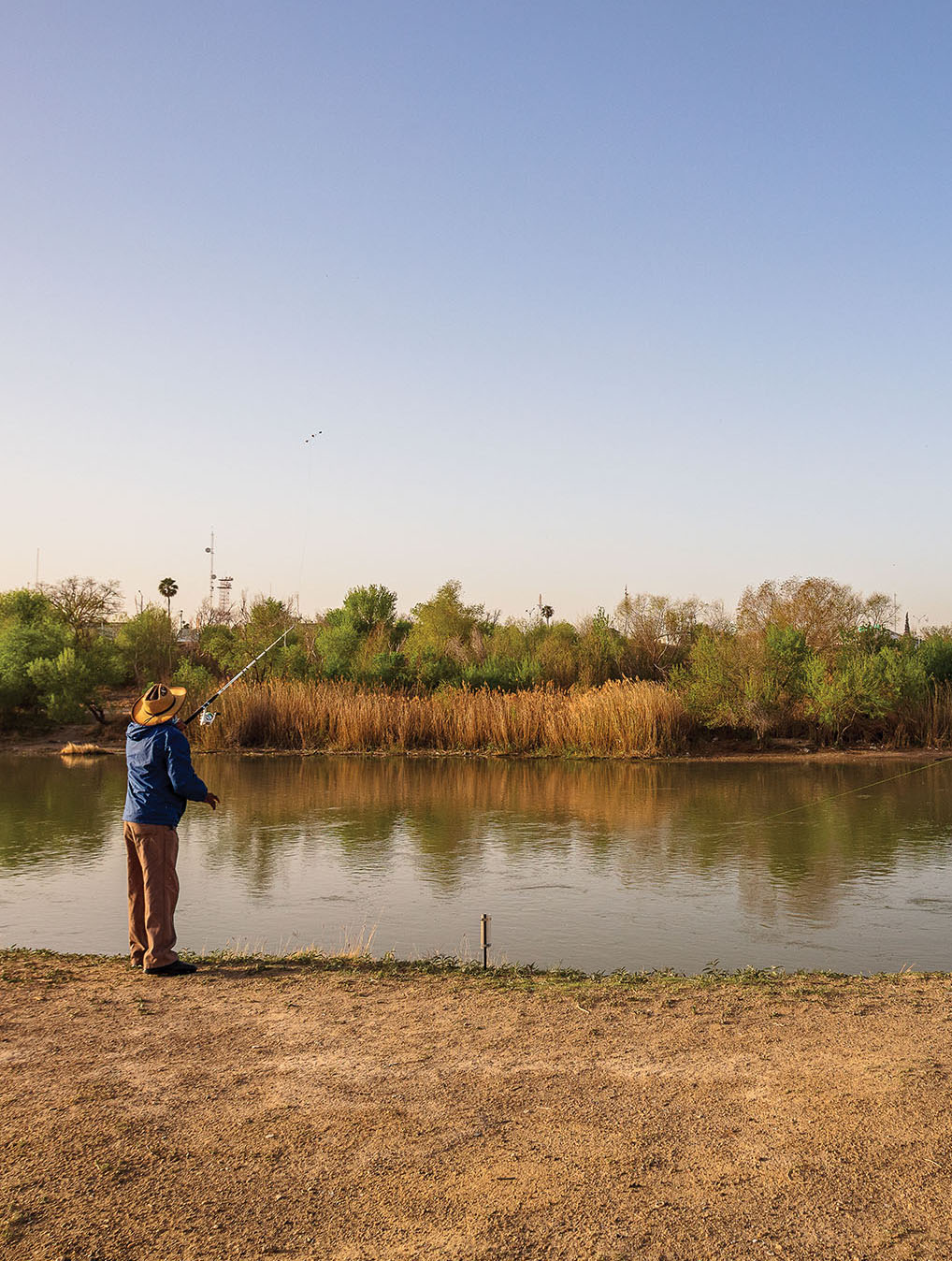 a picture of a man casting a fishing rod into gray brown water on a dirt shore