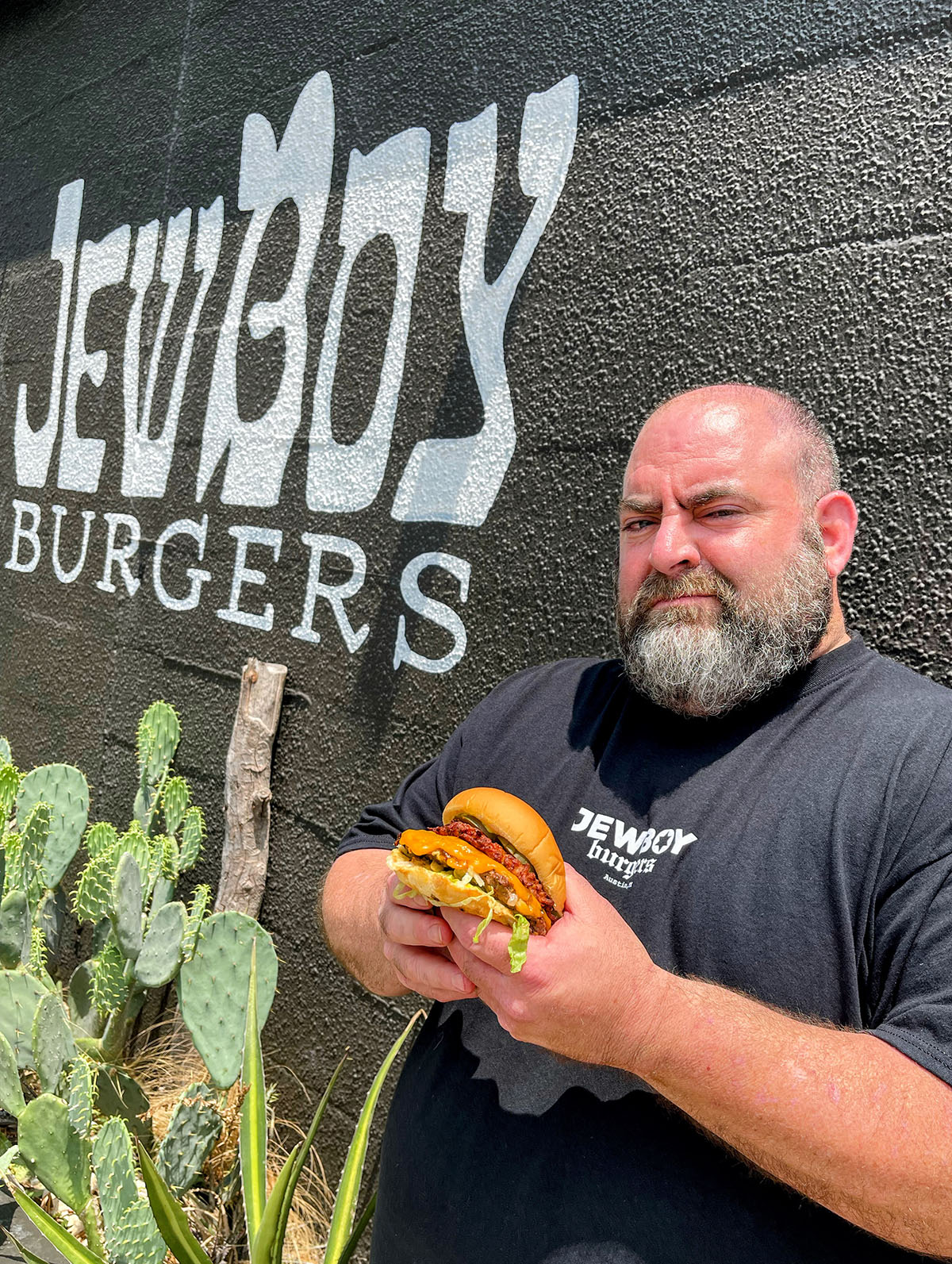 A man in a t-shirt holds a burger in front of a painted sign reading "JewBoy Burgers"