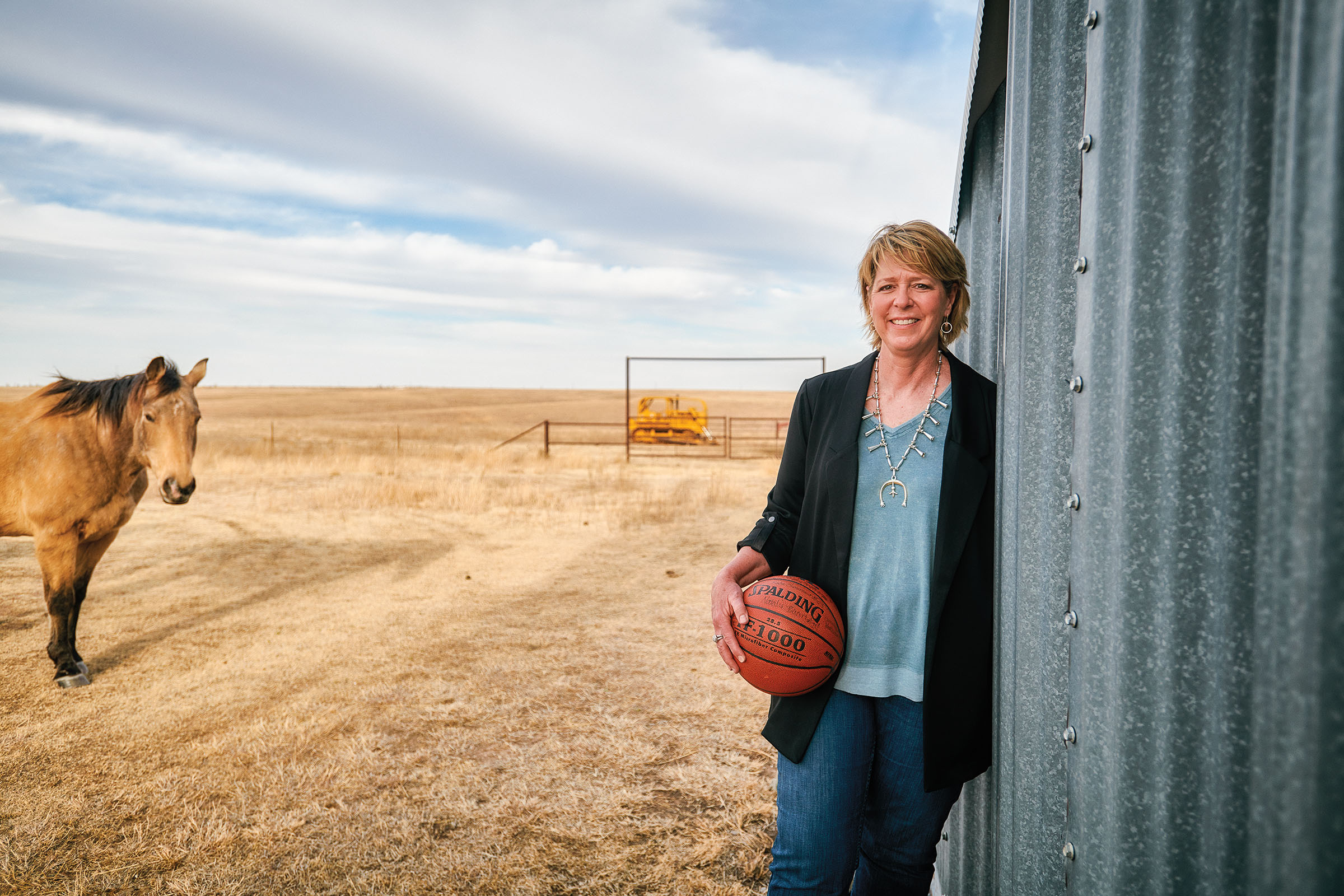 A woman in a blue shirt holds a basketball while leaning against a metal building on a farm