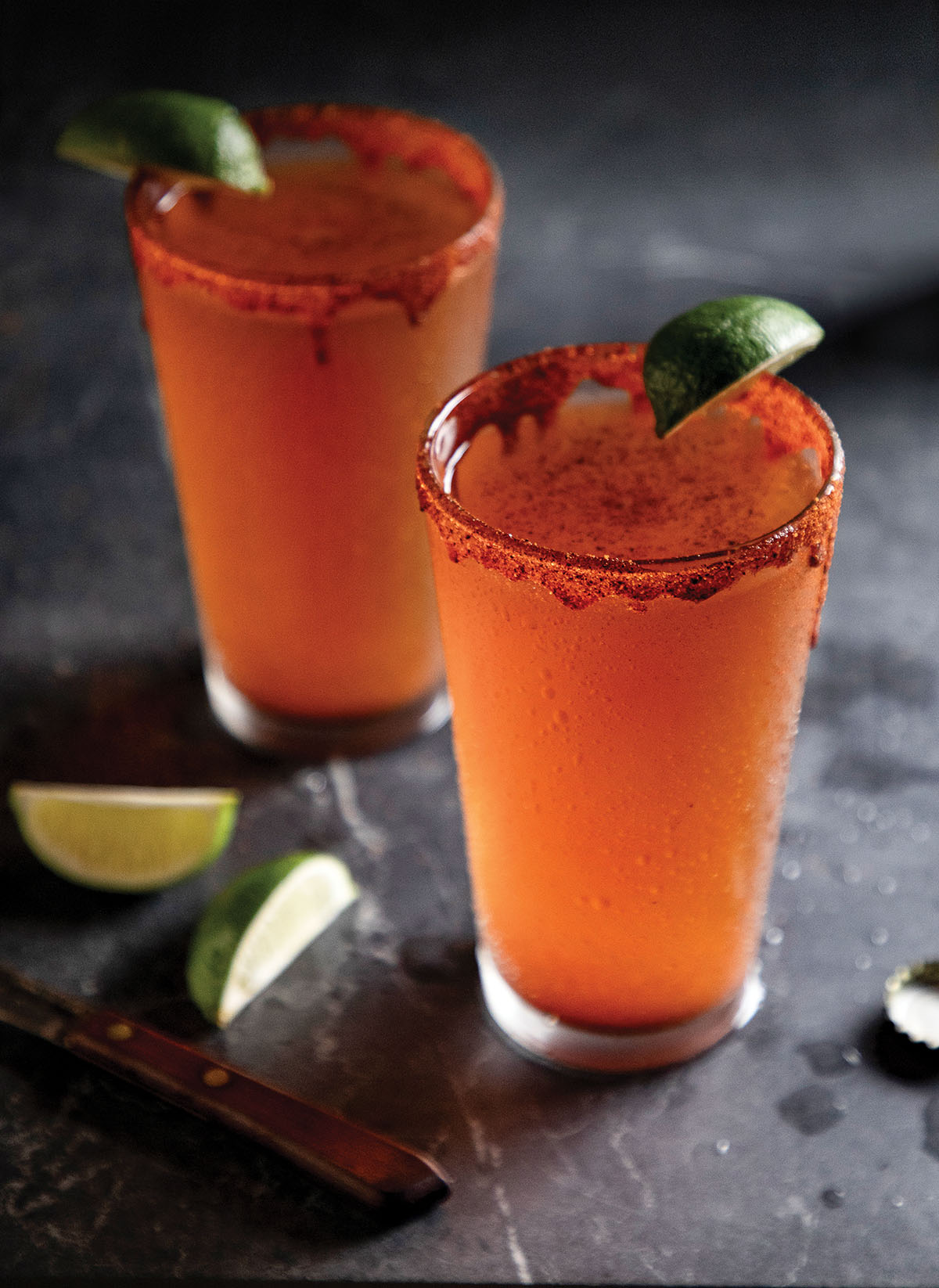 Two reddish-brown glasses of michelada with a lime and chile salt on the rim