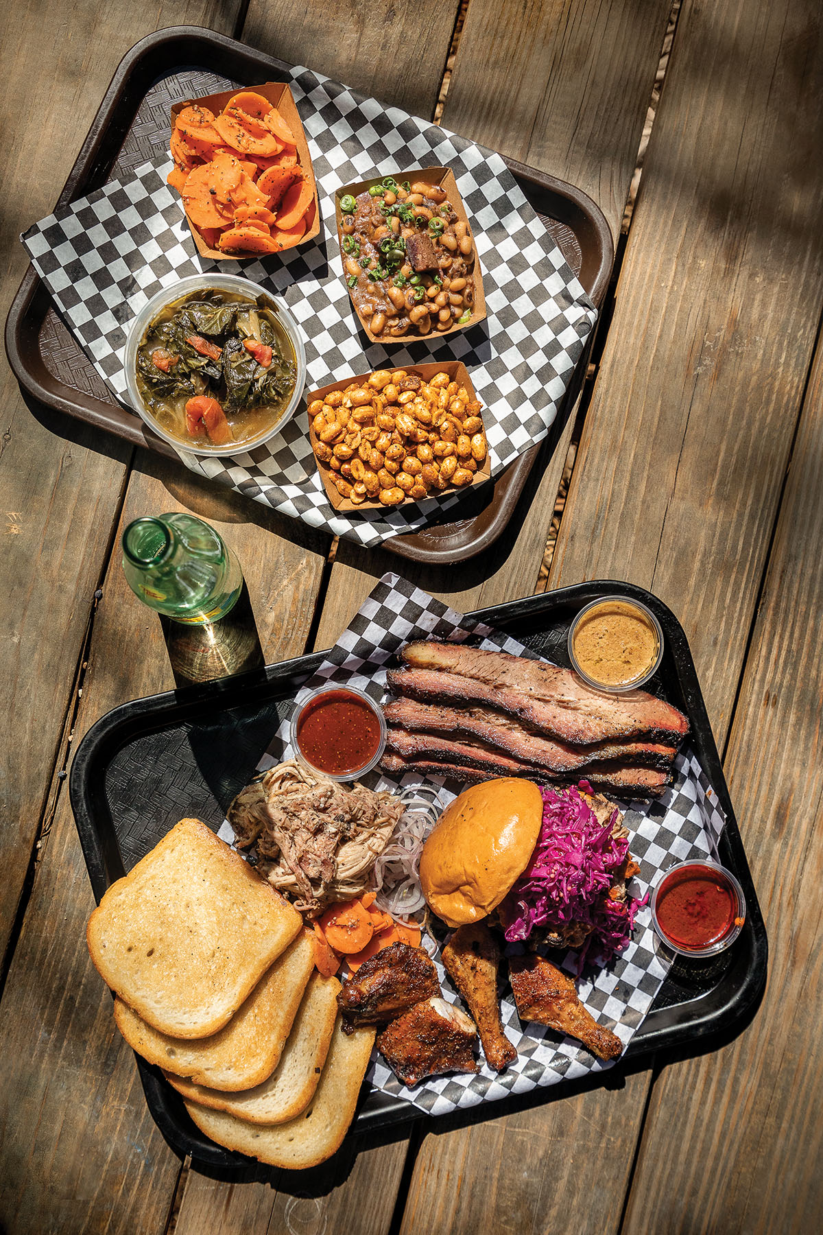 An overhead view of two trays of barbecued meats, bread, side dishes on a checkered paper backing