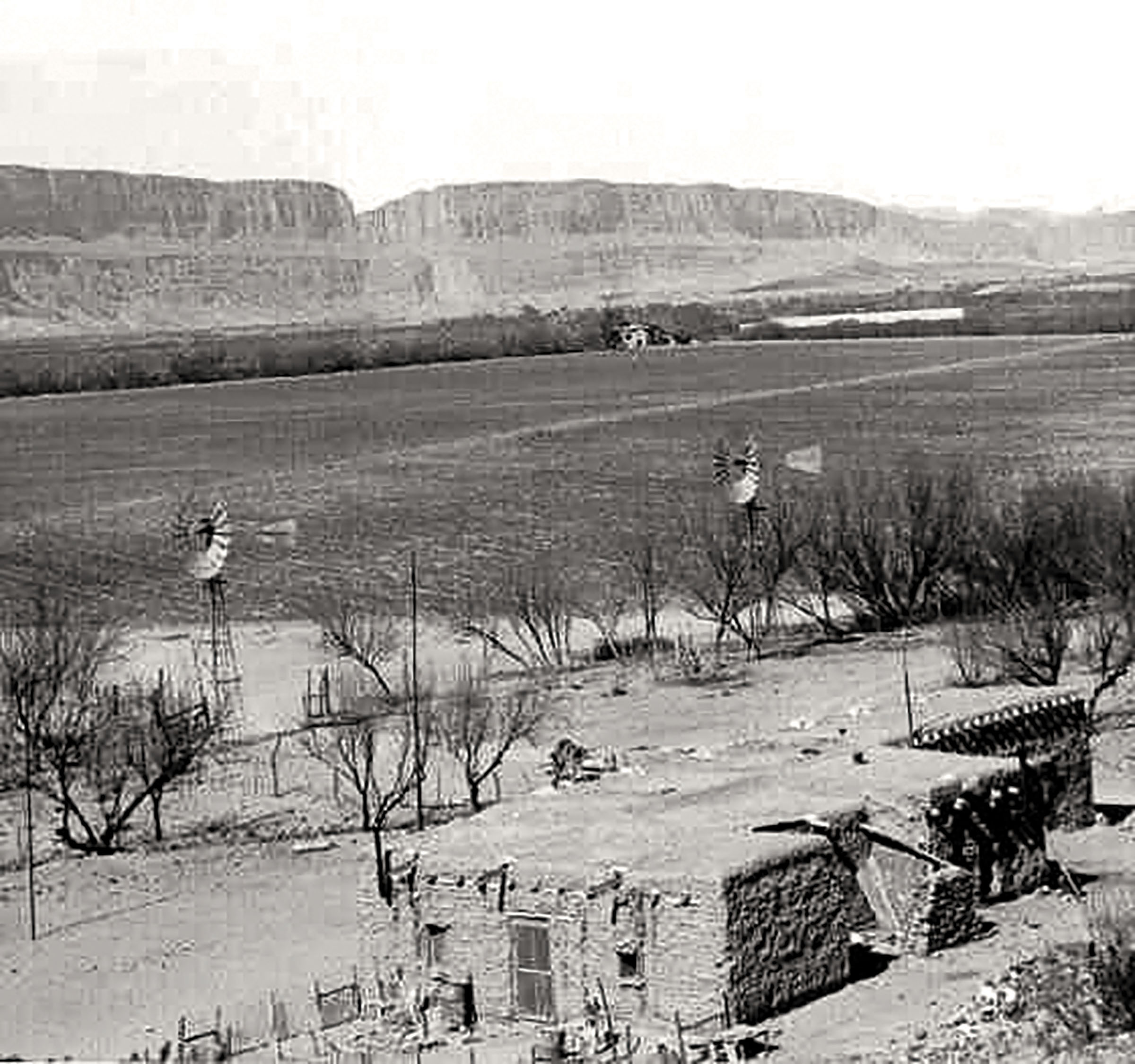 A black and white photo of a humble adobe home along the water beneath tall mountains in the distance