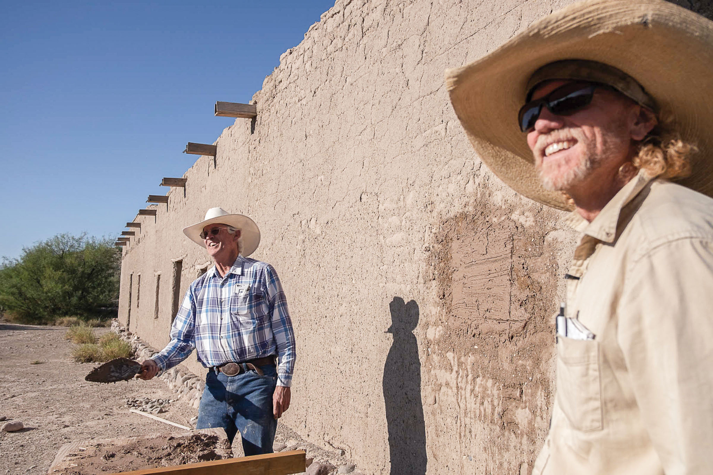 Two men in hats look into the sun in front of a patched, darker portion of an adobe wall