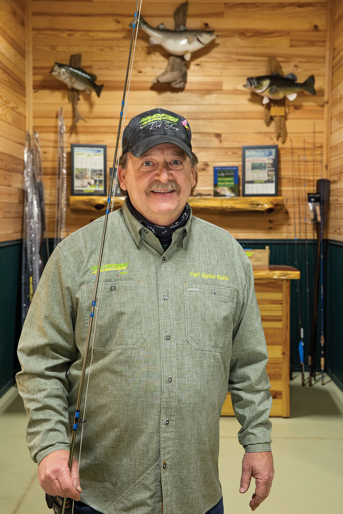 A man in a green shirt and baseball cap stands holding a fishing rod in front of a wood-paneled wall