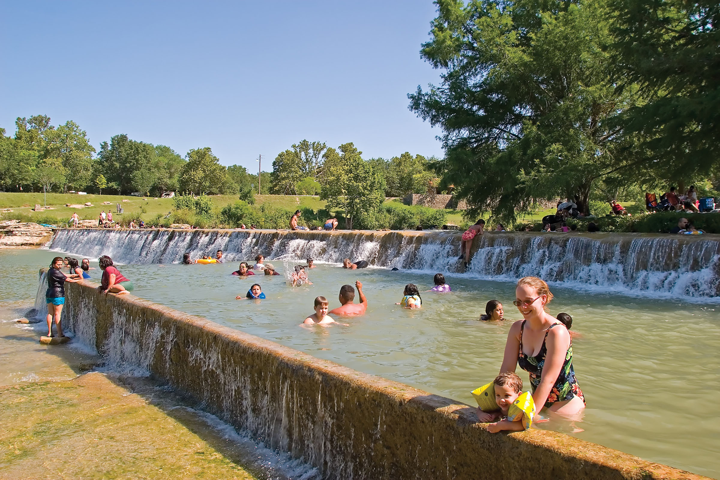 A large group of people float and swim in rushing water at Blanco State Park