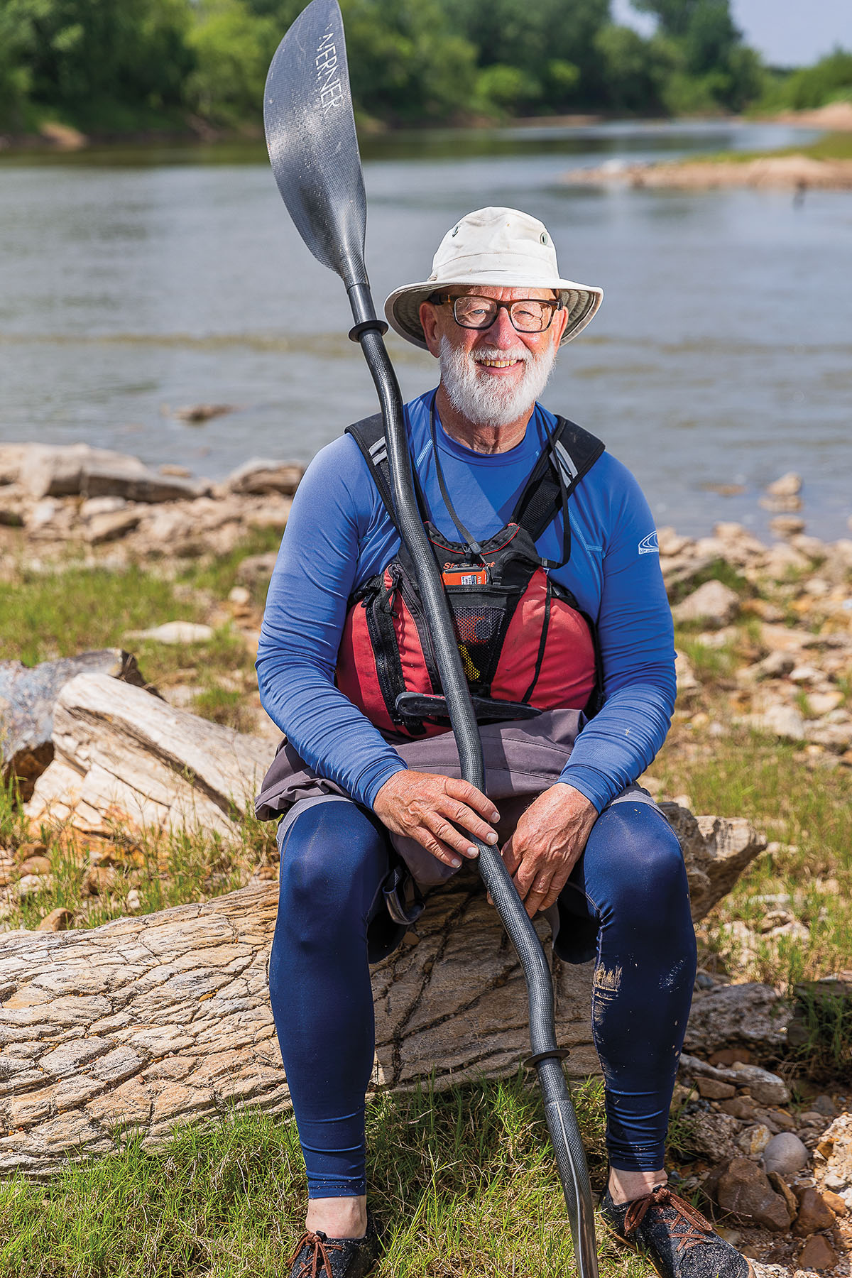 A man in a white hat and wearing a PFD holds a large paddle on the banks of the Brazos