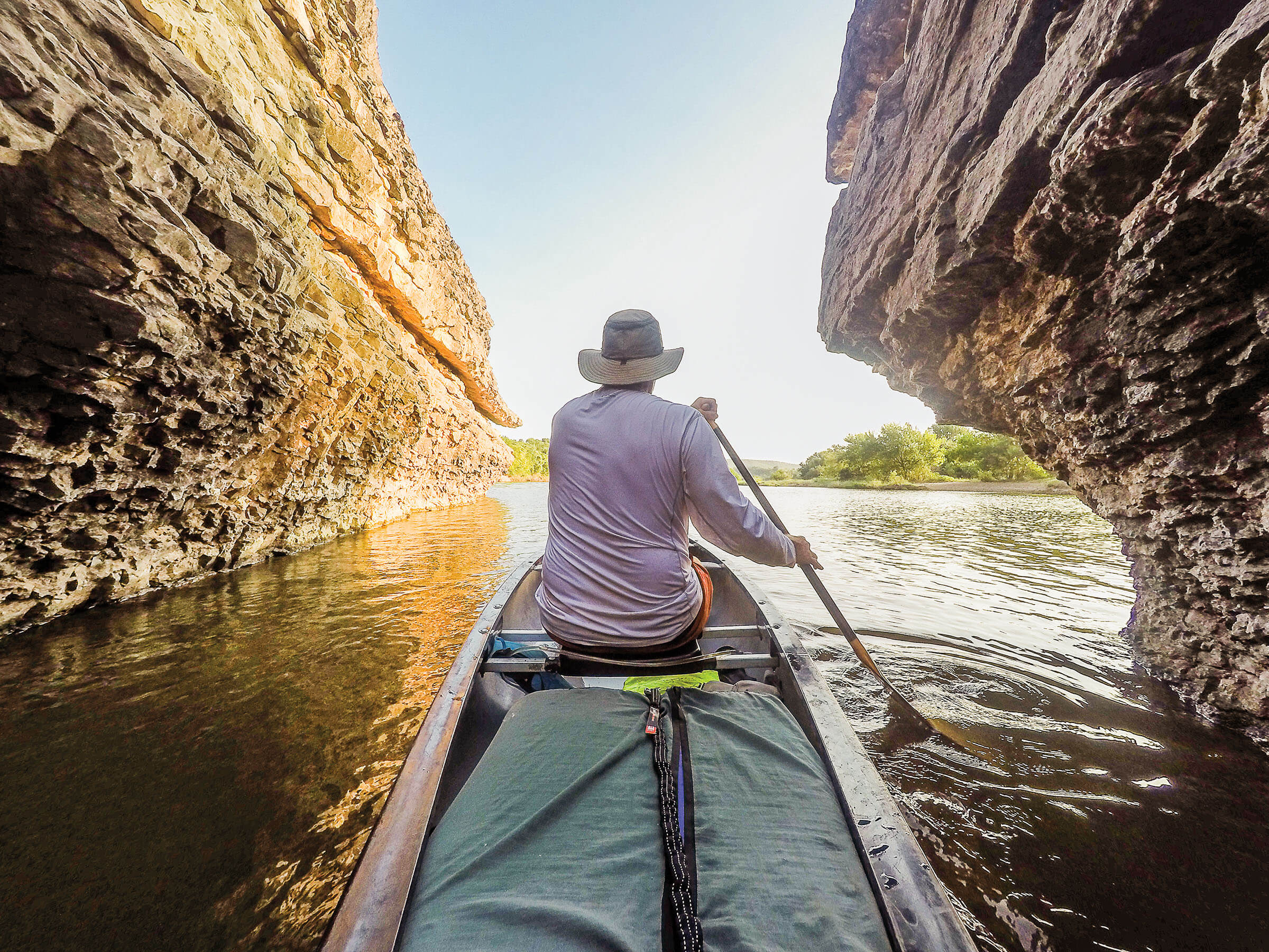 A man in a long-sleeved white shirt paddles between two rock outcroppings on a river