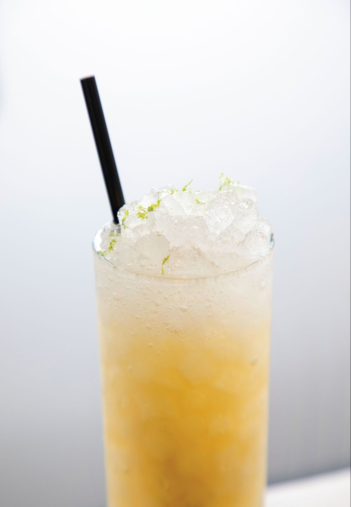 A golden-colored cocktail in a tall glass with ice, lime garnish and a black straw