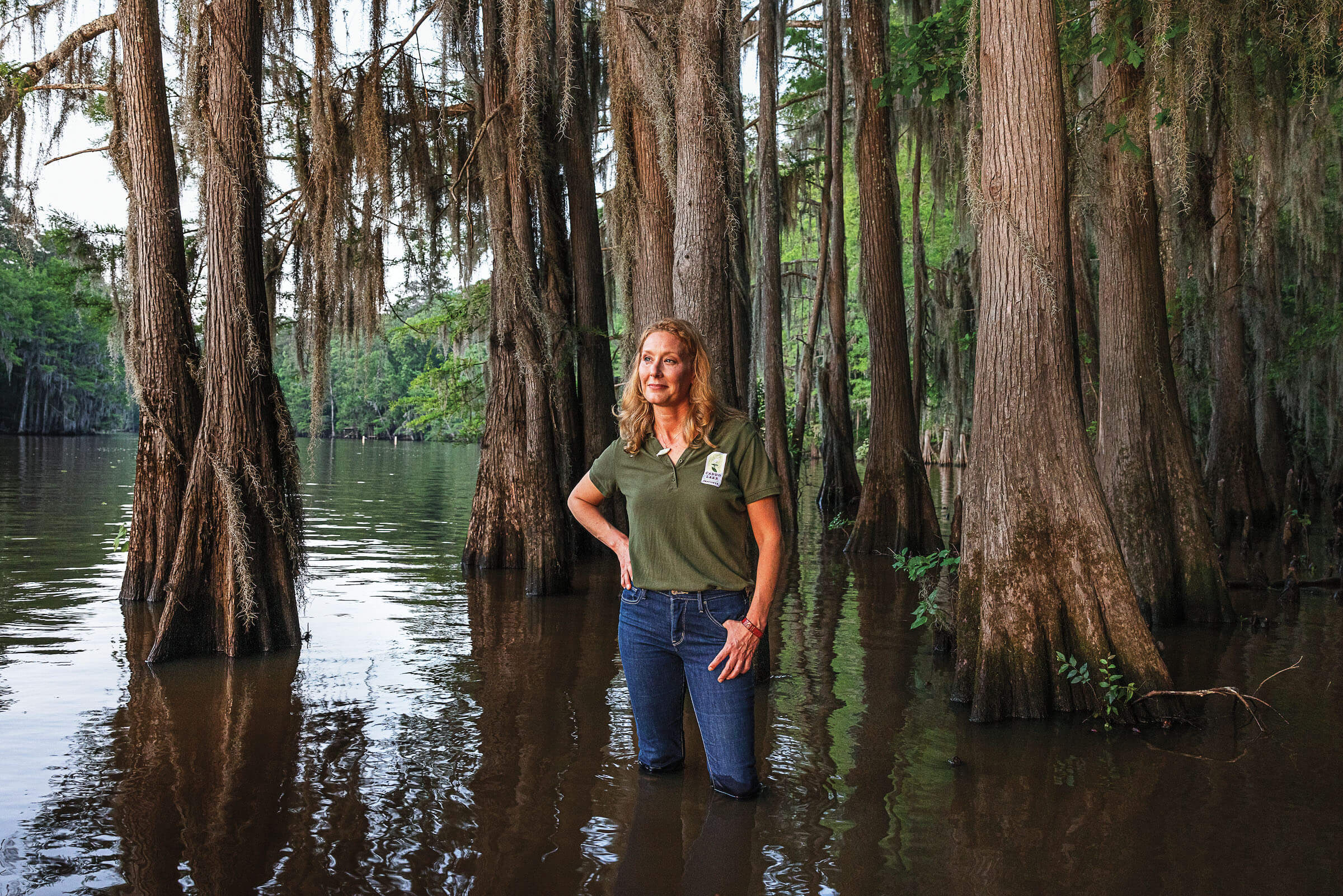 A person in a green shirt and jeans stands in the water beneath large trees