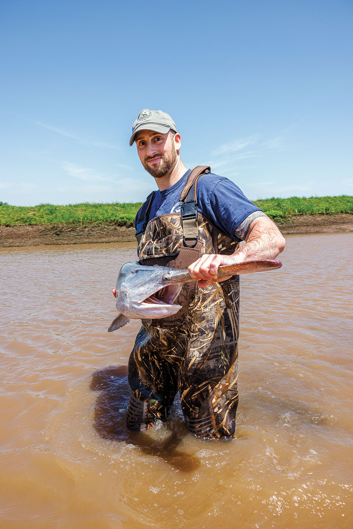 A man in camoflauge hip waders holds a paddlefish in brown water