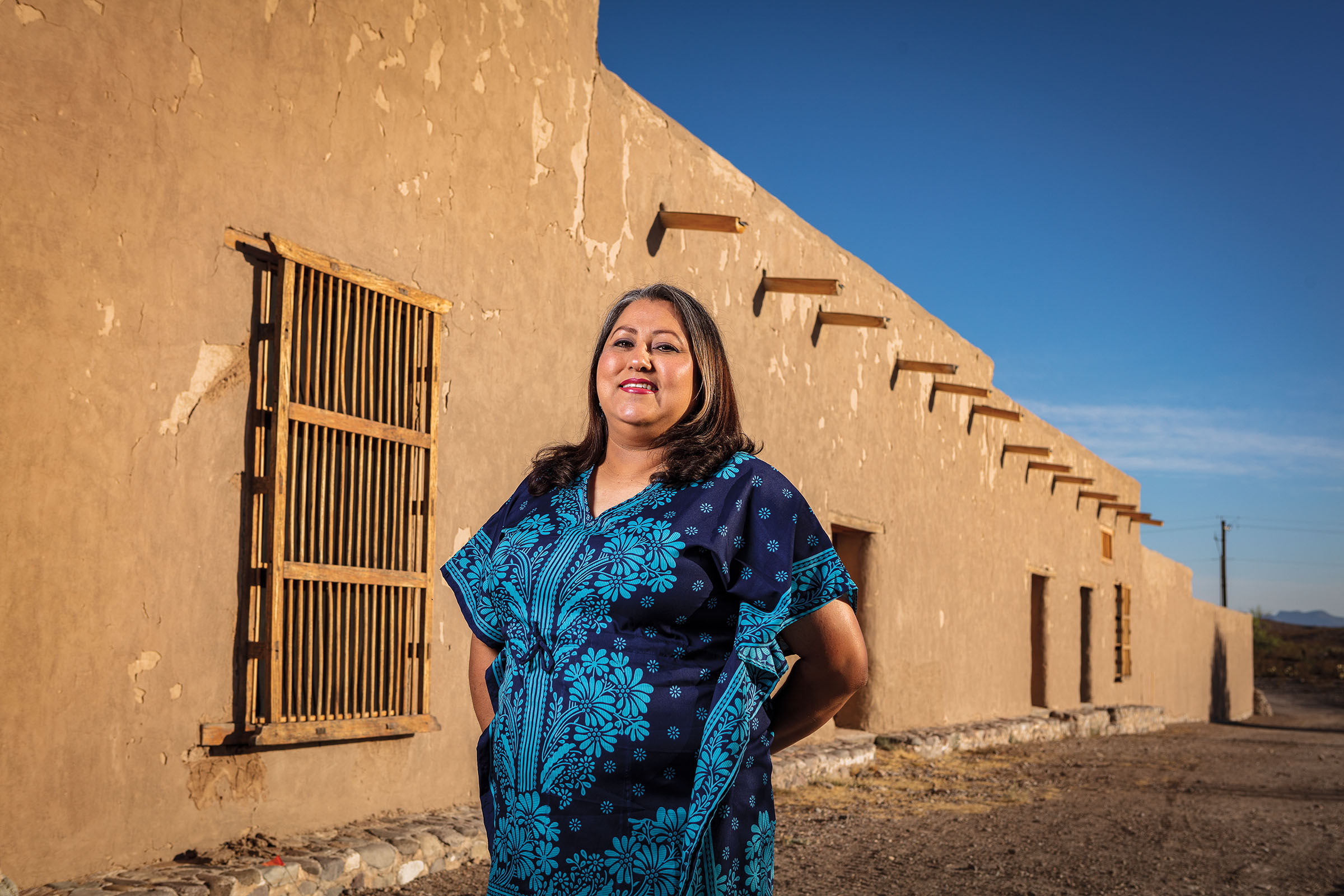 A woman stands outside of an adobe building under a clear blue sky