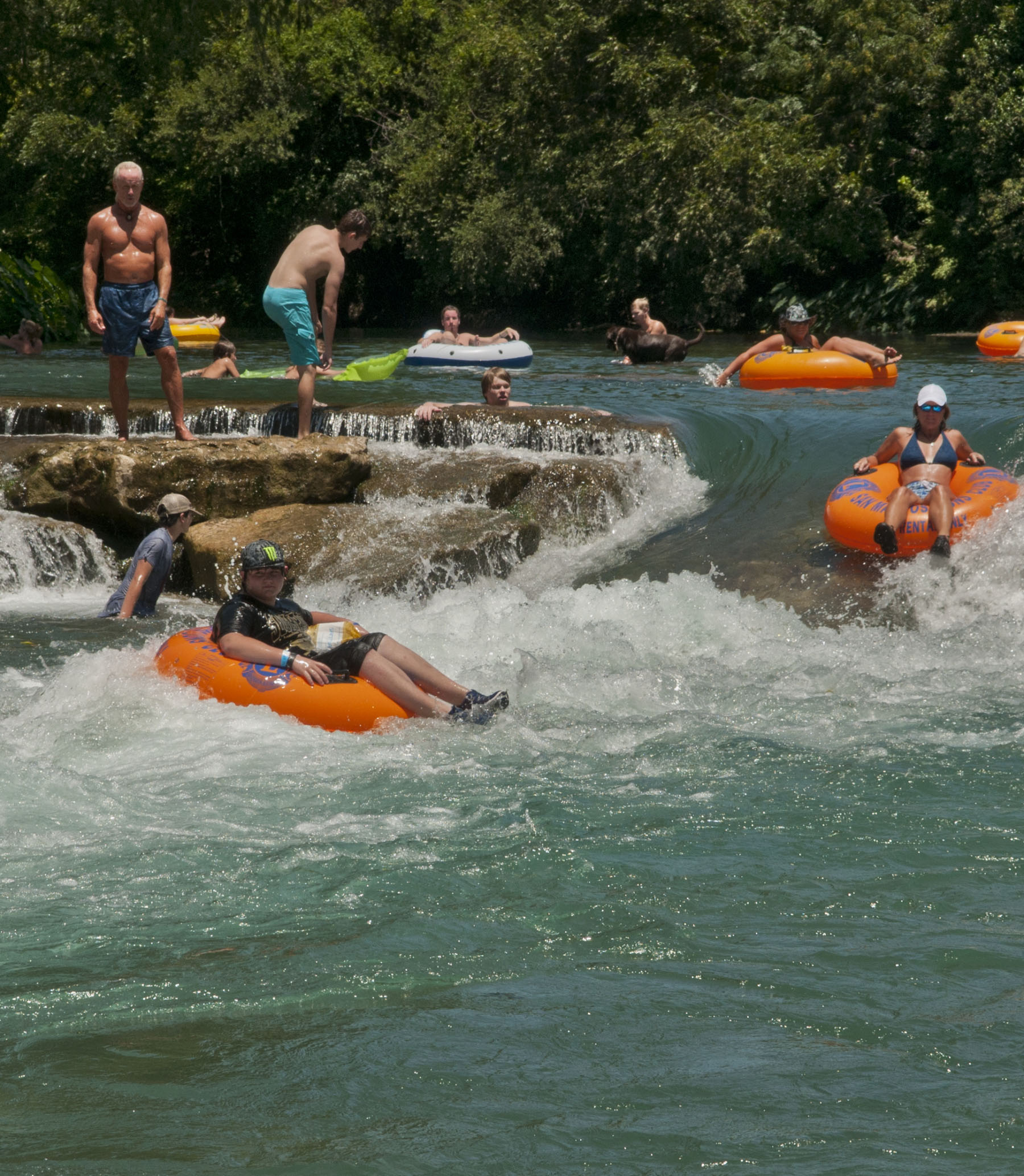 A group of people float down a small rapid in bright orange tubes in blue green water