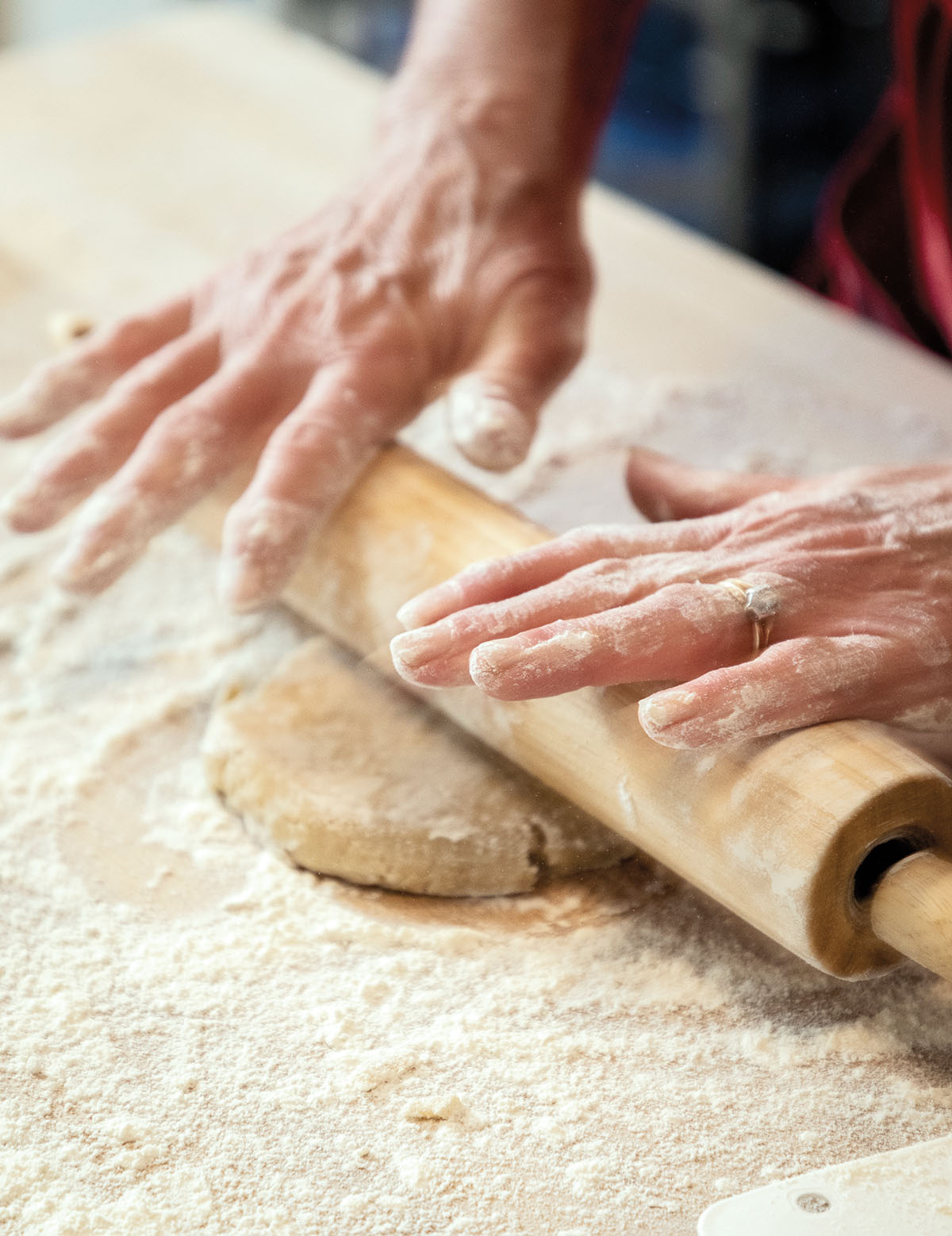 A person's flour-covered hands push a wooden rolling pin over a block of brown dough