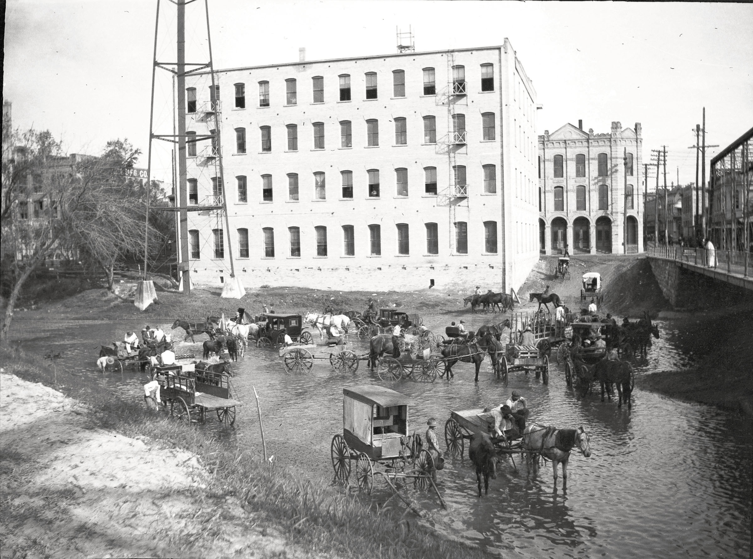 A large group of horses and buggies move through the water in front of a white building in a vintage photograph