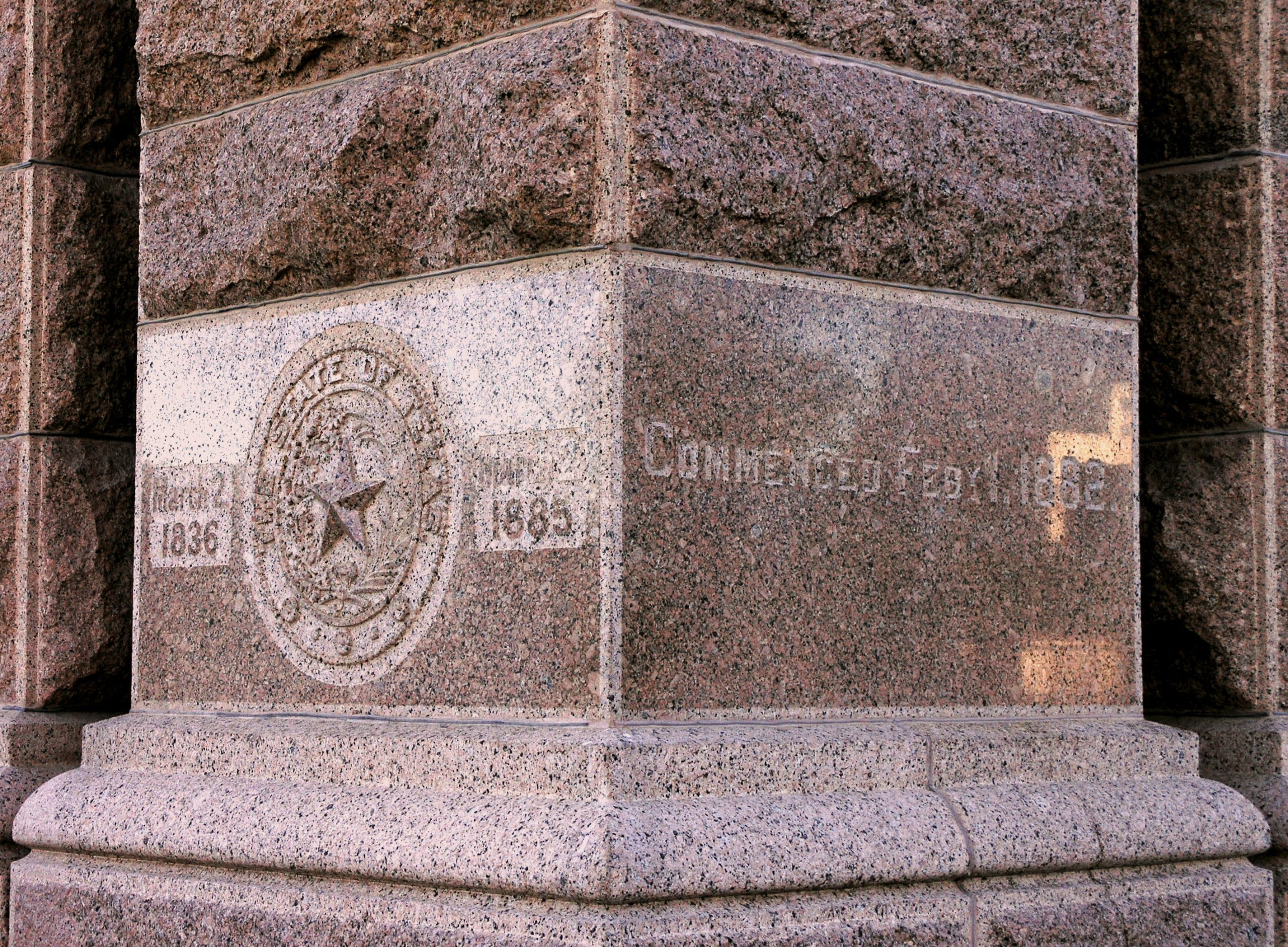 A smooth red granite block at the base of a column with the seal of the state of Texas engraved into it, and the years 1836 and 1885.